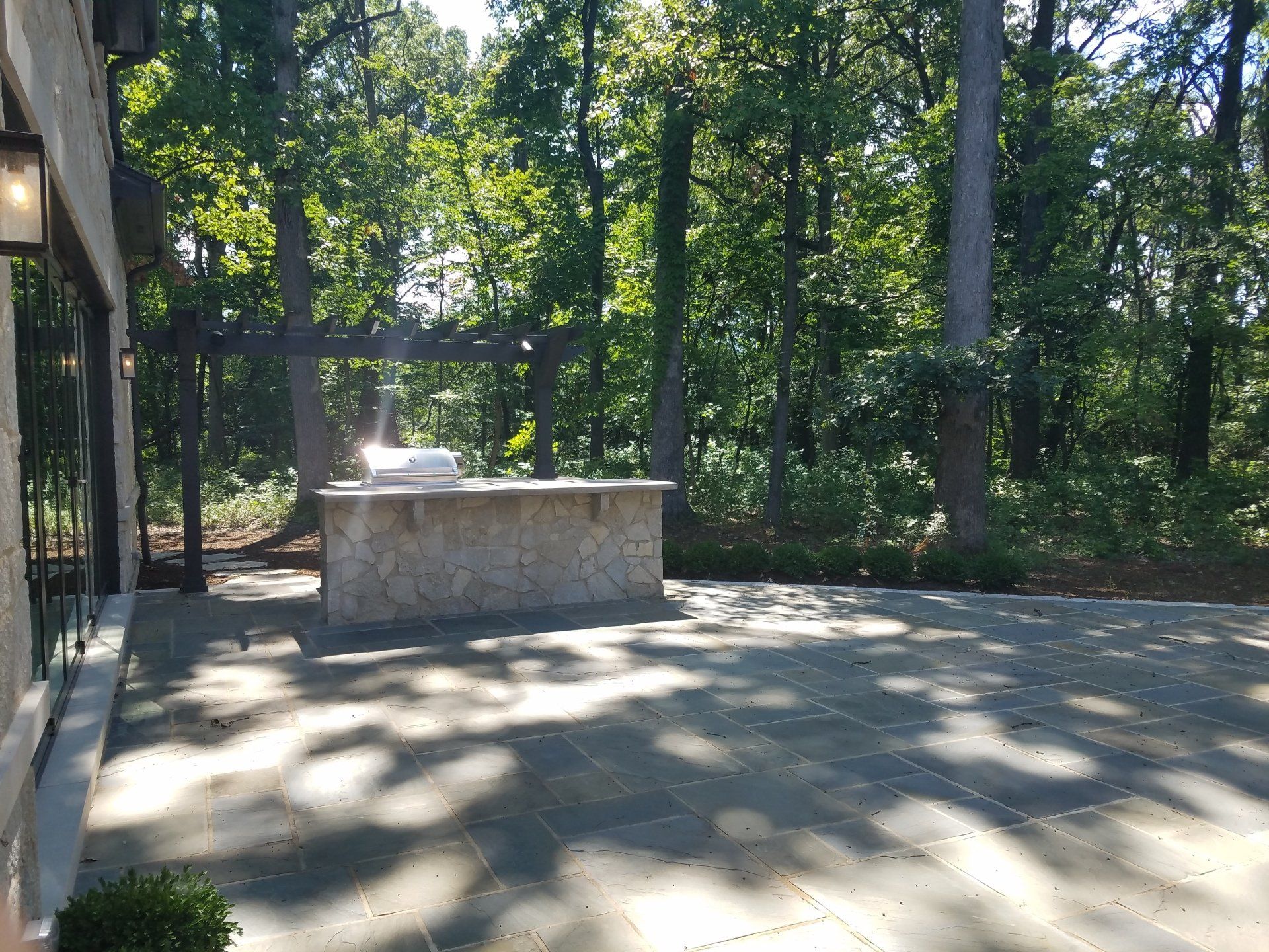 Stone patio with built-in grill, surrounded by trees and a partially visible building.