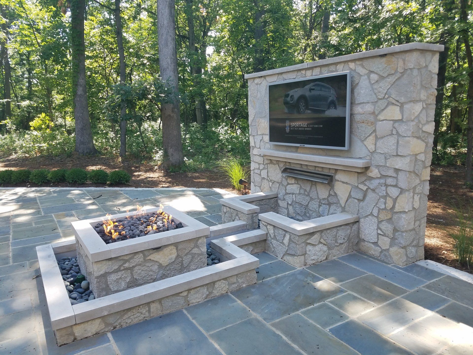 Stone patio with fire pit and outdoor TV on a stacked stone wall in a wooded setting.