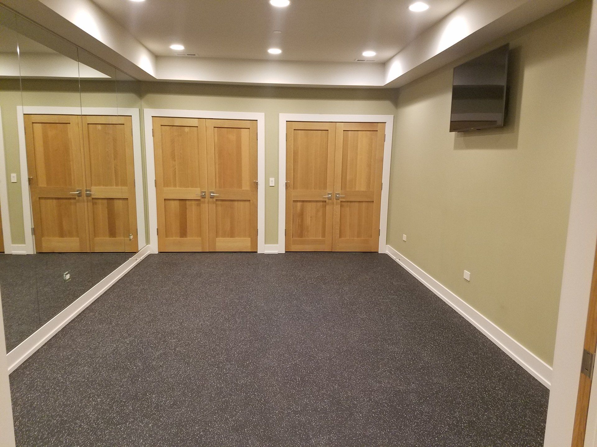A room with wood doors, a dark speckled floor, and a mirror on the left wall. A TV is mounted on the right.