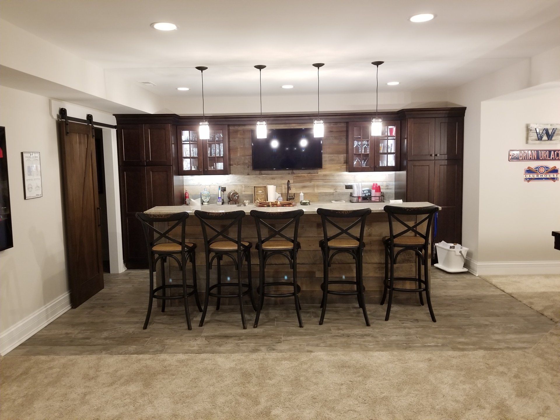 Basement bar with wood cabinetry, bar stools, and a barn door, lit by pendant lights.