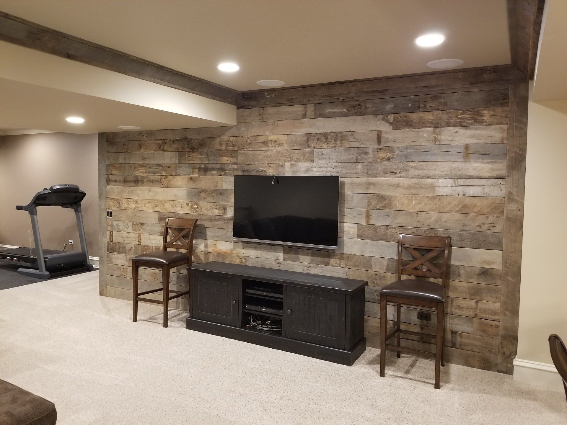 A home entertainment area with a TV on a rustic wood-paneled wall, a treadmill, and two wooden bar stools.