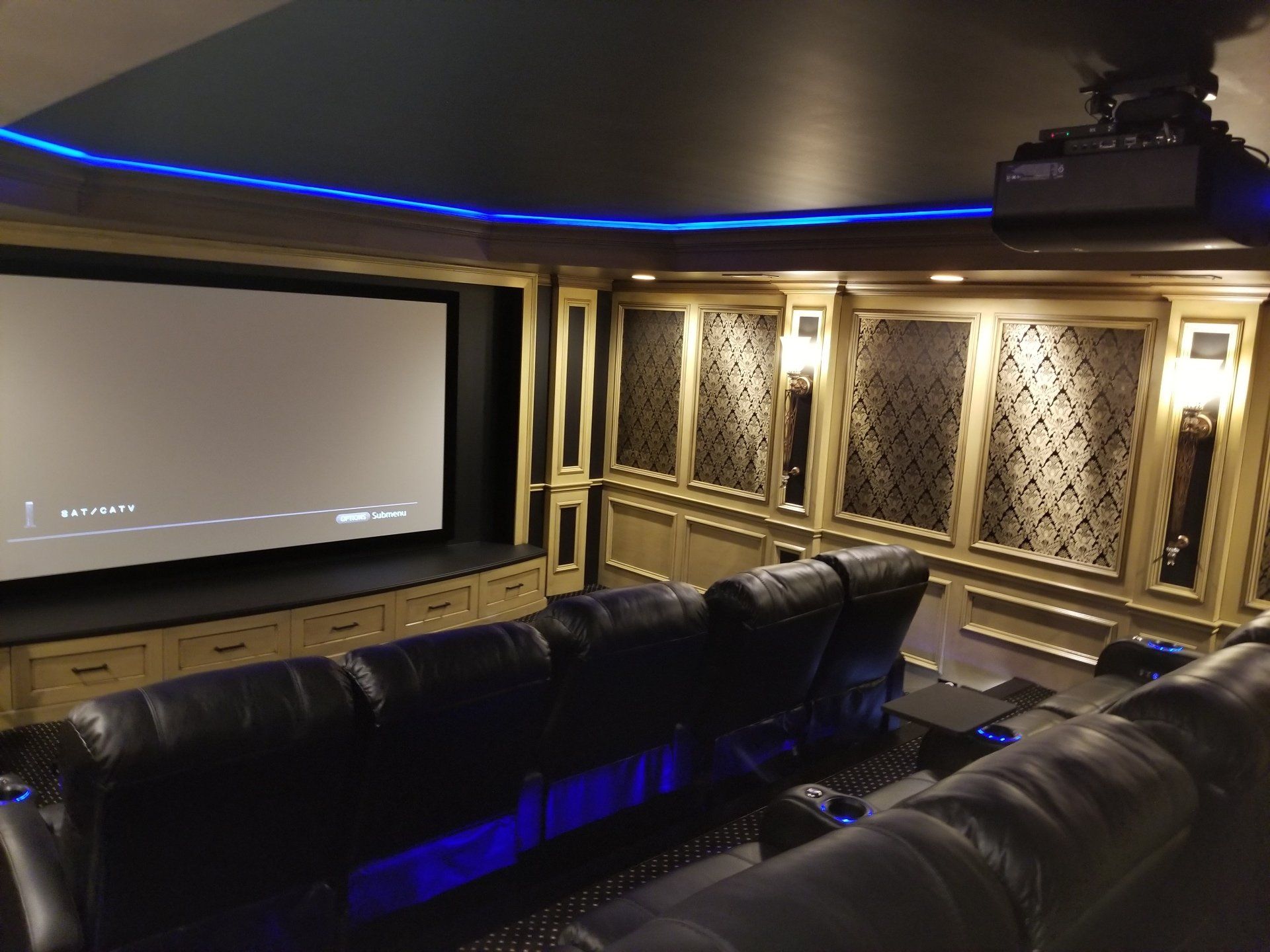 Home theater with black leather seating, large screen, and ornate wall panels, lit with blue and gold accents.