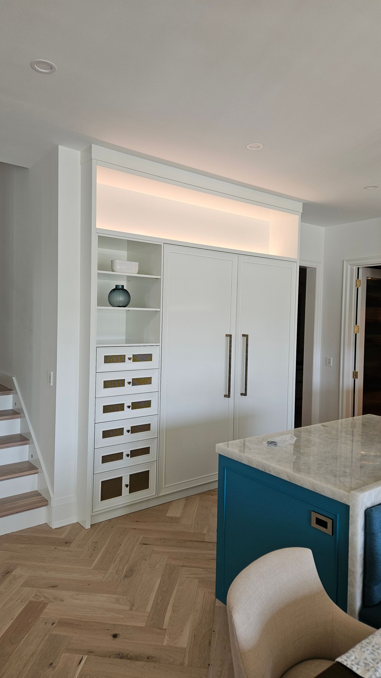 White kitchen with built-in fridge, blue island, wooden floor, and a staircase.
