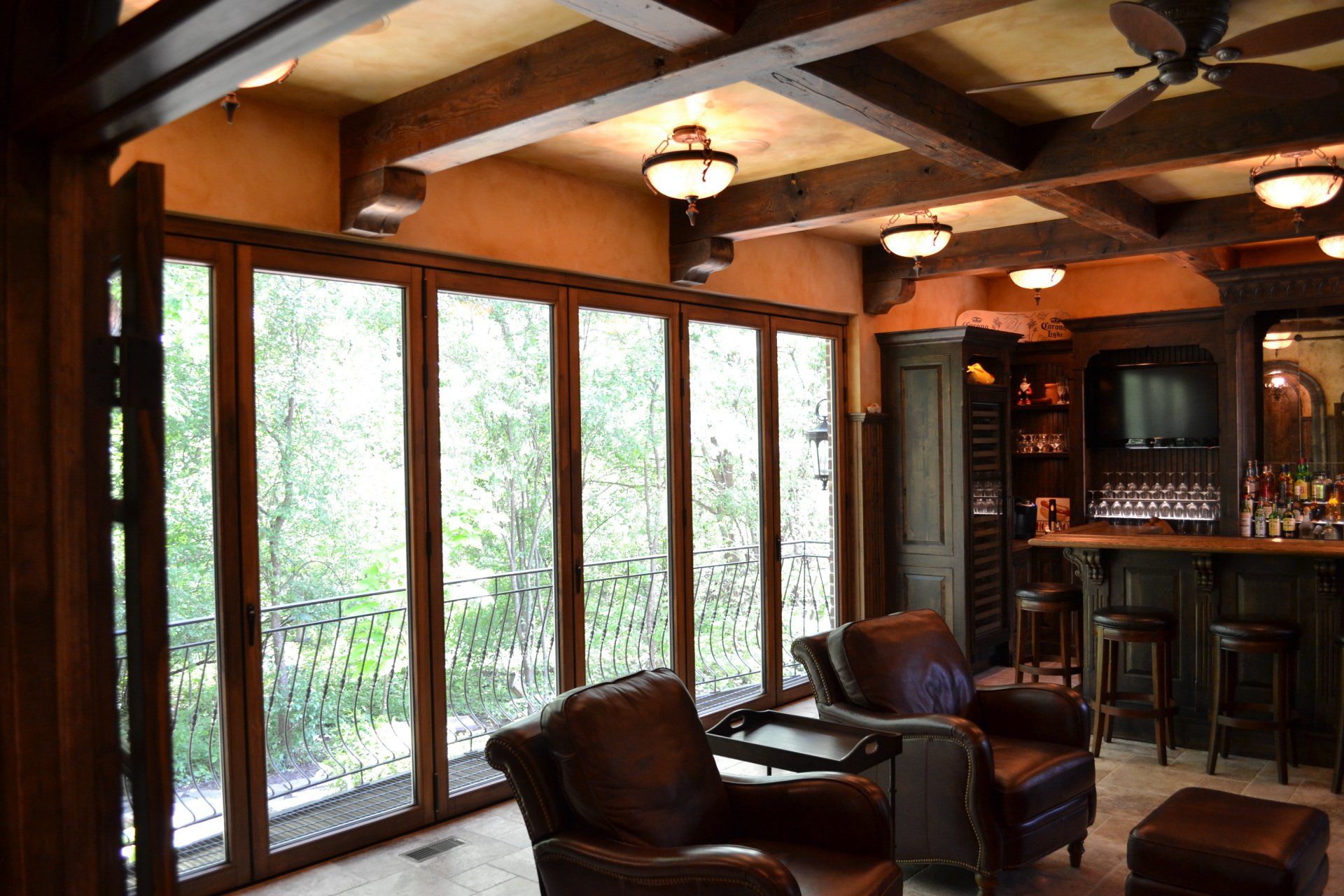 Living room with folding glass doors, wooden beams, bar, leather chairs, and view of trees.
