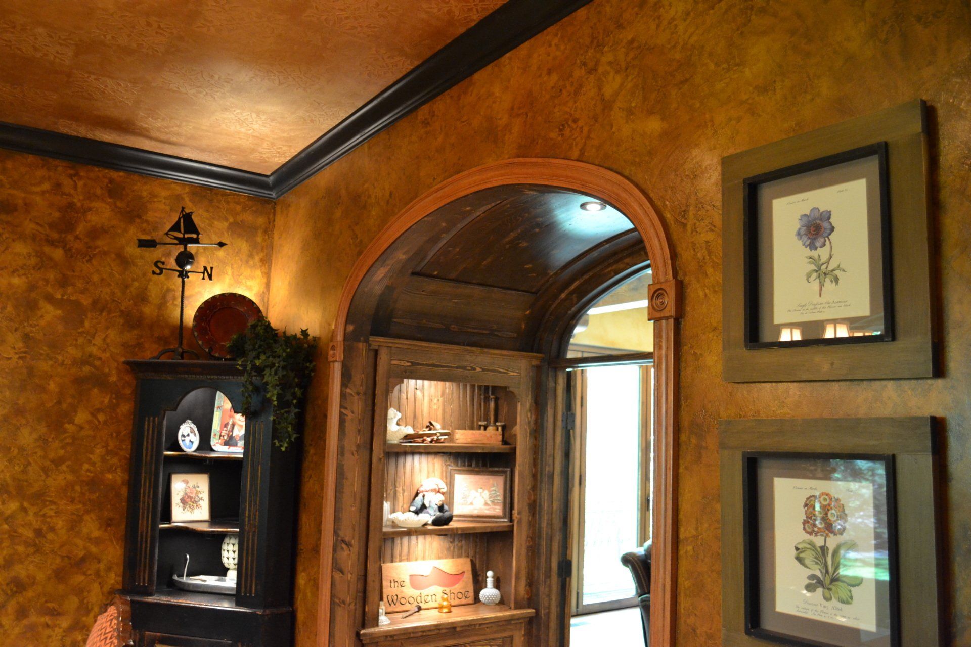 Corner of a room with ornate walls, a corner cabinet, arched doorway, and framed botanical prints.