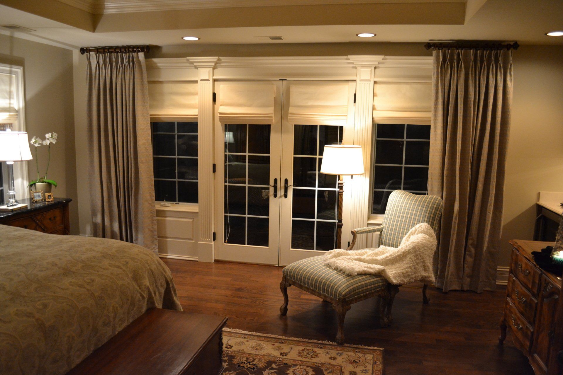 Bedroom with dark wood floor, French doors, beige curtains, and a chaise lounge.
