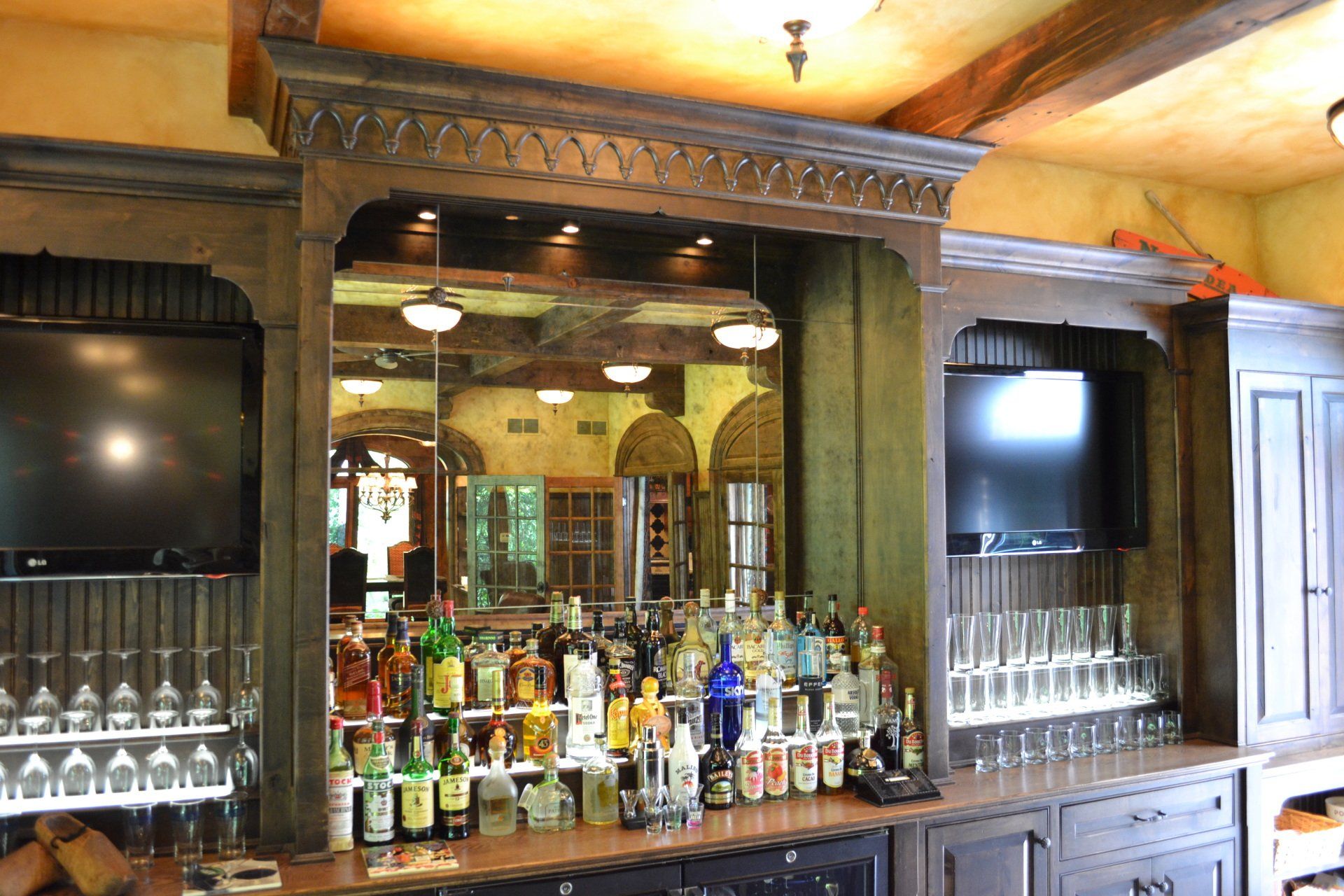 Bar with bottles, glassware, and TVs in a room with a mirror.