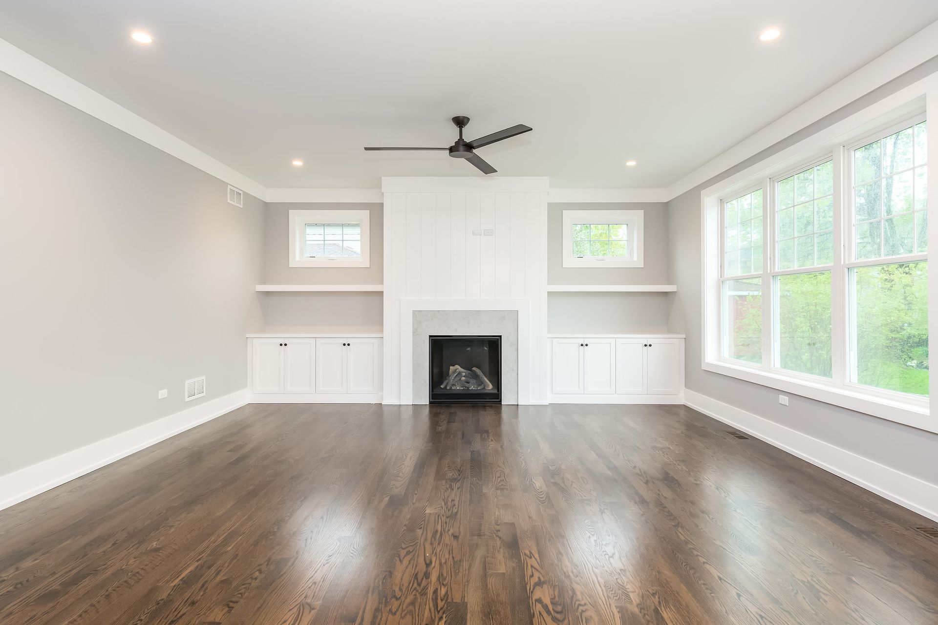 Empty living room with fireplace, built-ins, dark wood floor, and large windows.