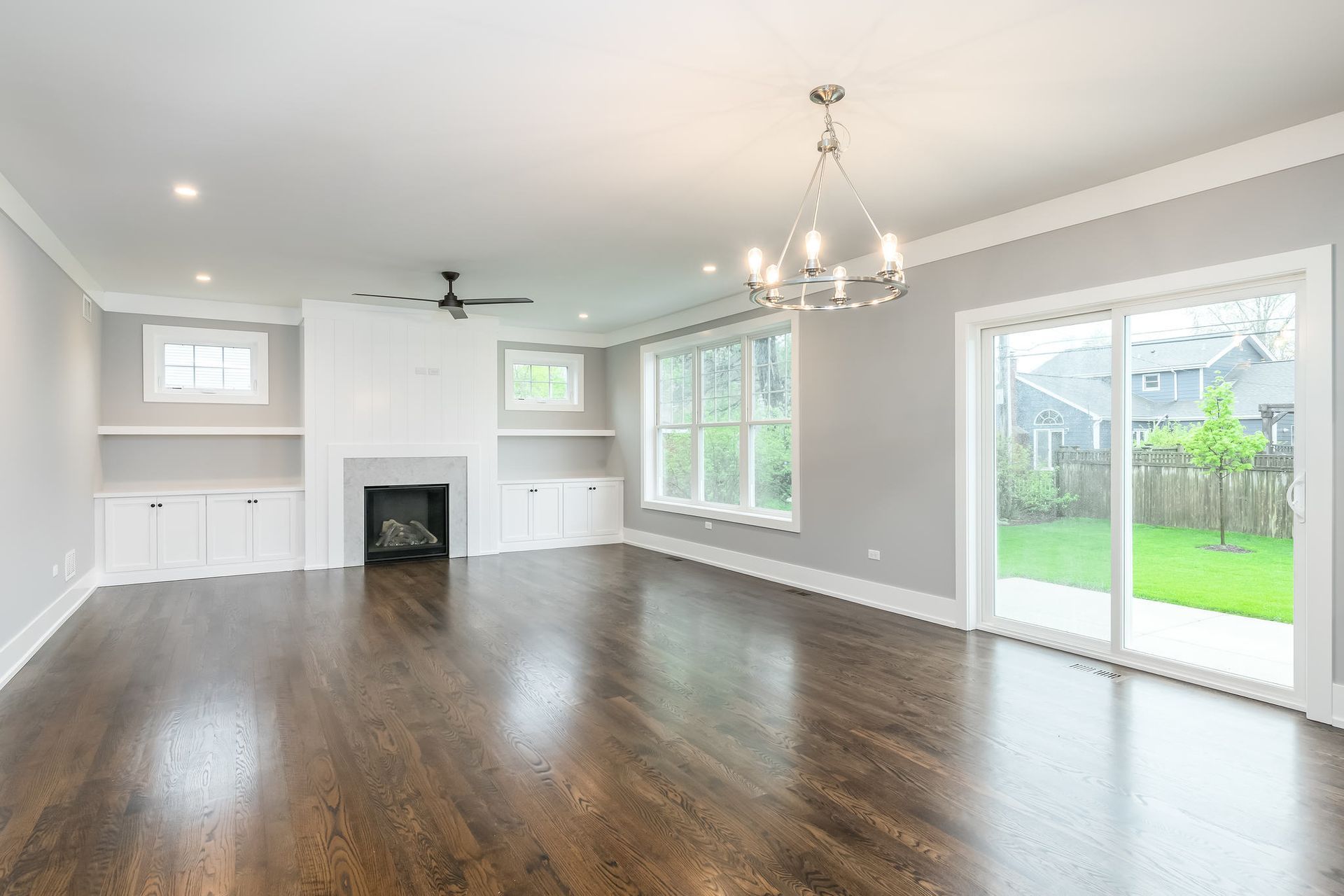 Empty living room with dark wood floors, white trim, fireplace, and large windows.