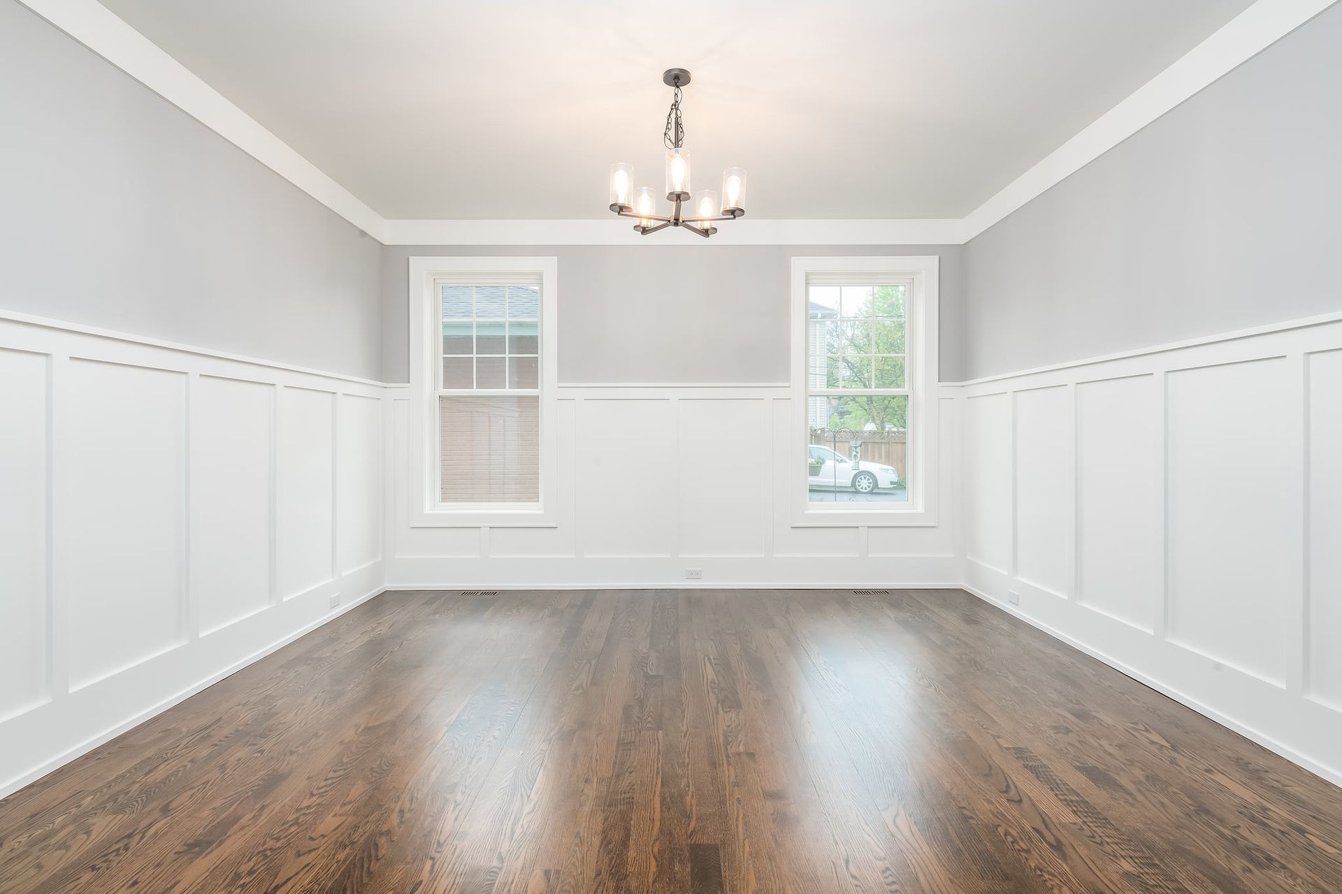 Empty, light-gray dining room with white wainscoting, two windows, and a dark hardwood floor.