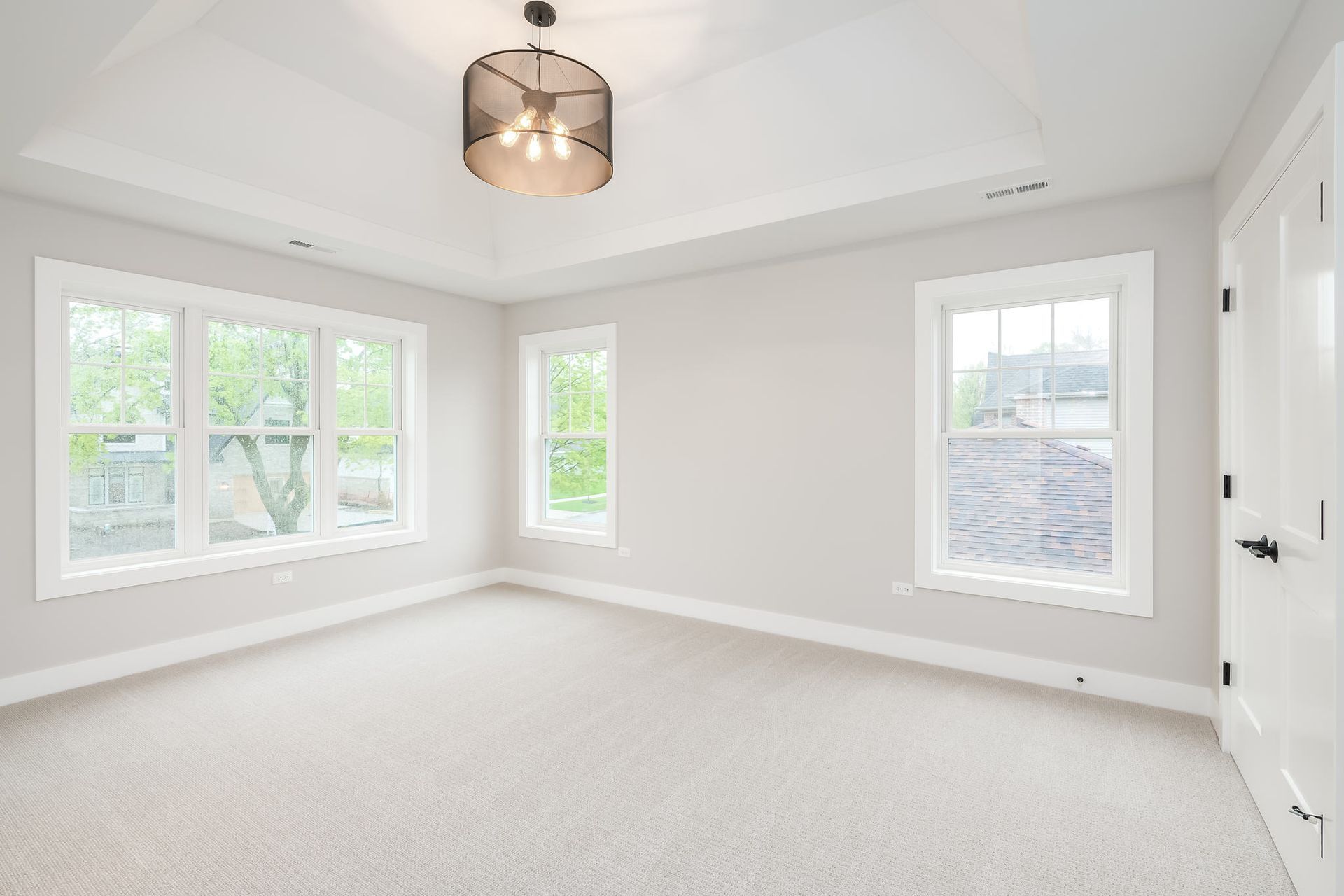 Empty, light-filled bedroom with three windows, white trim, and light gray carpet. Decorative ceiling light.
