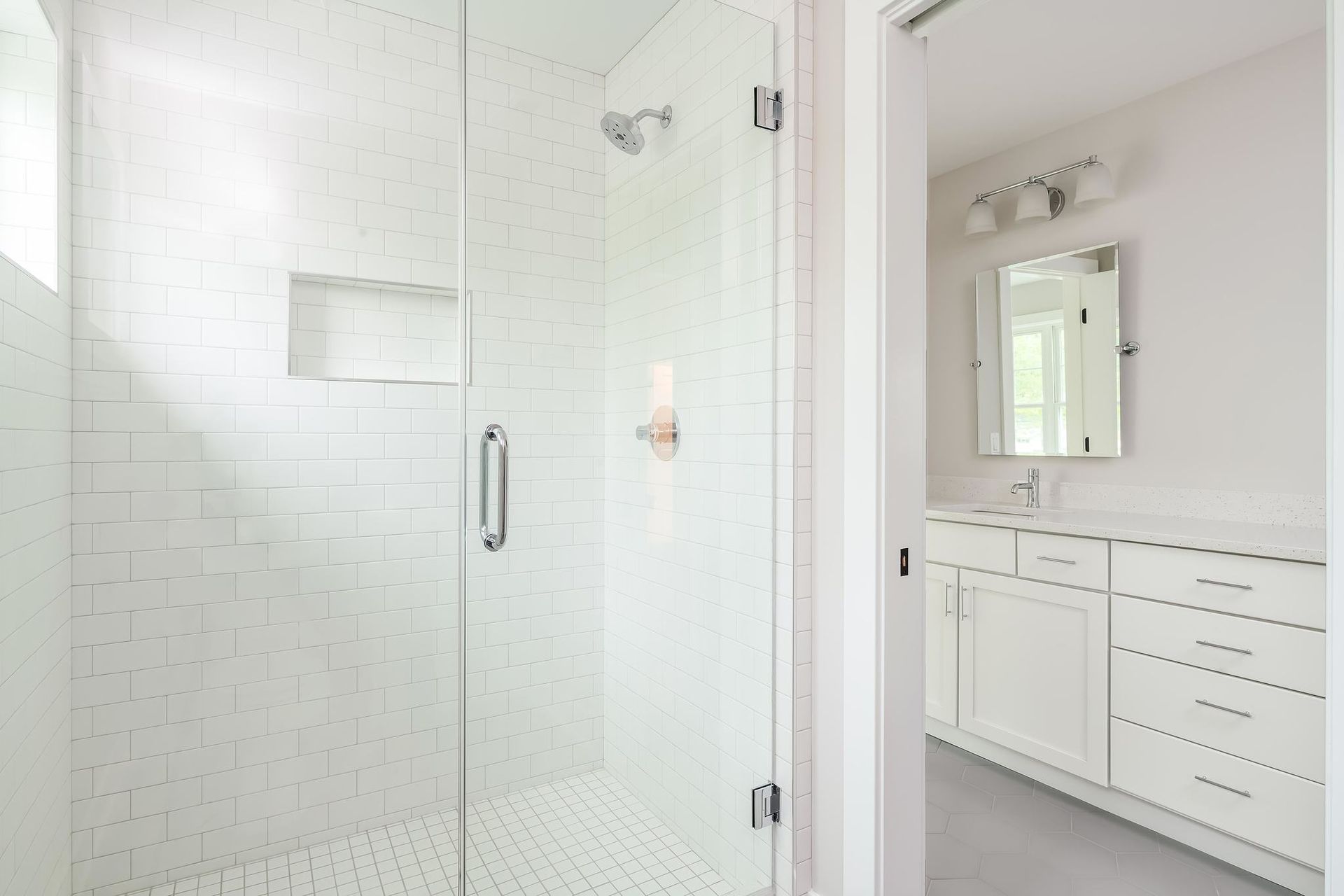 White-tiled shower with glass door, opening to a bathroom with a sink, vanity, and mirror.