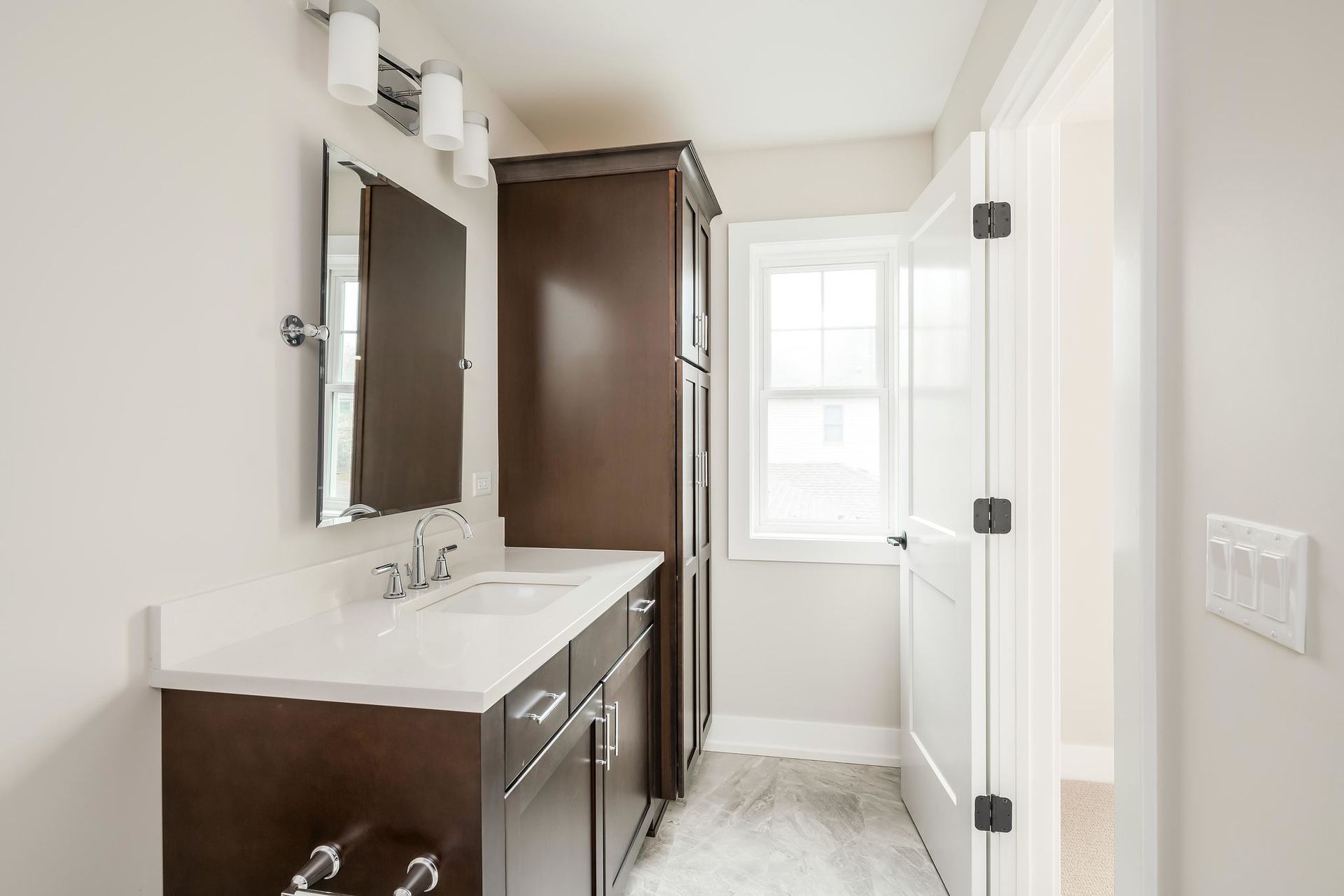 Bathroom with dark brown vanity, tall cabinet, small window, and open door.