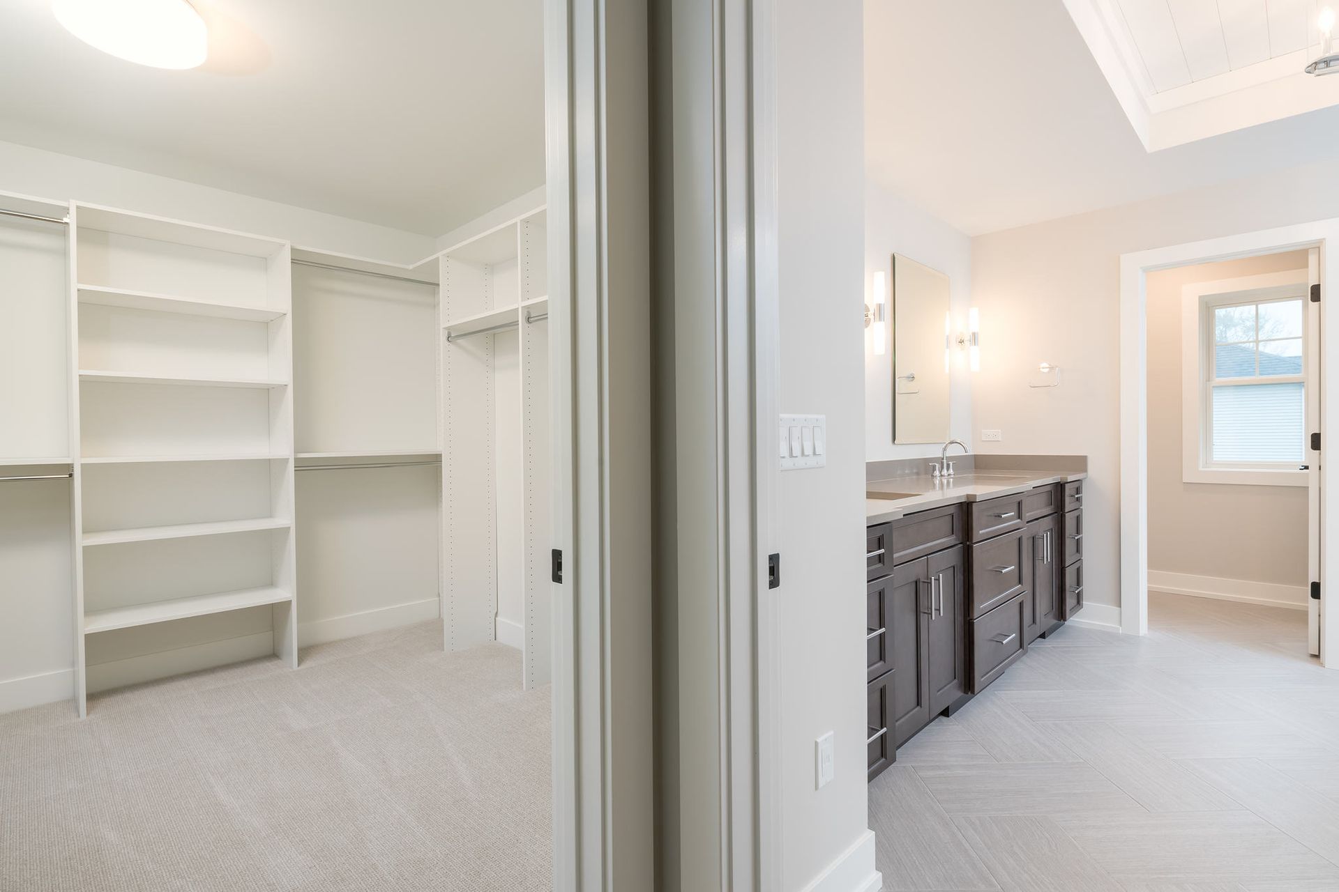 Walk-in closet and bathroom with white shelving and dark brown vanity. Soft grey carpet.