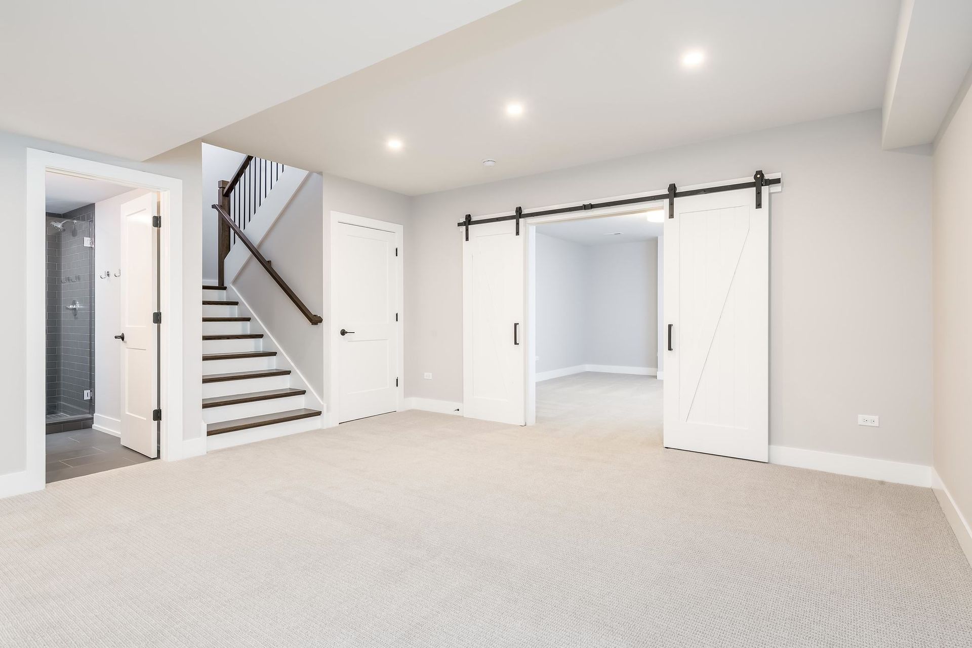 Empty, light-colored basement room with carpet, stairs, and sliding white doors.