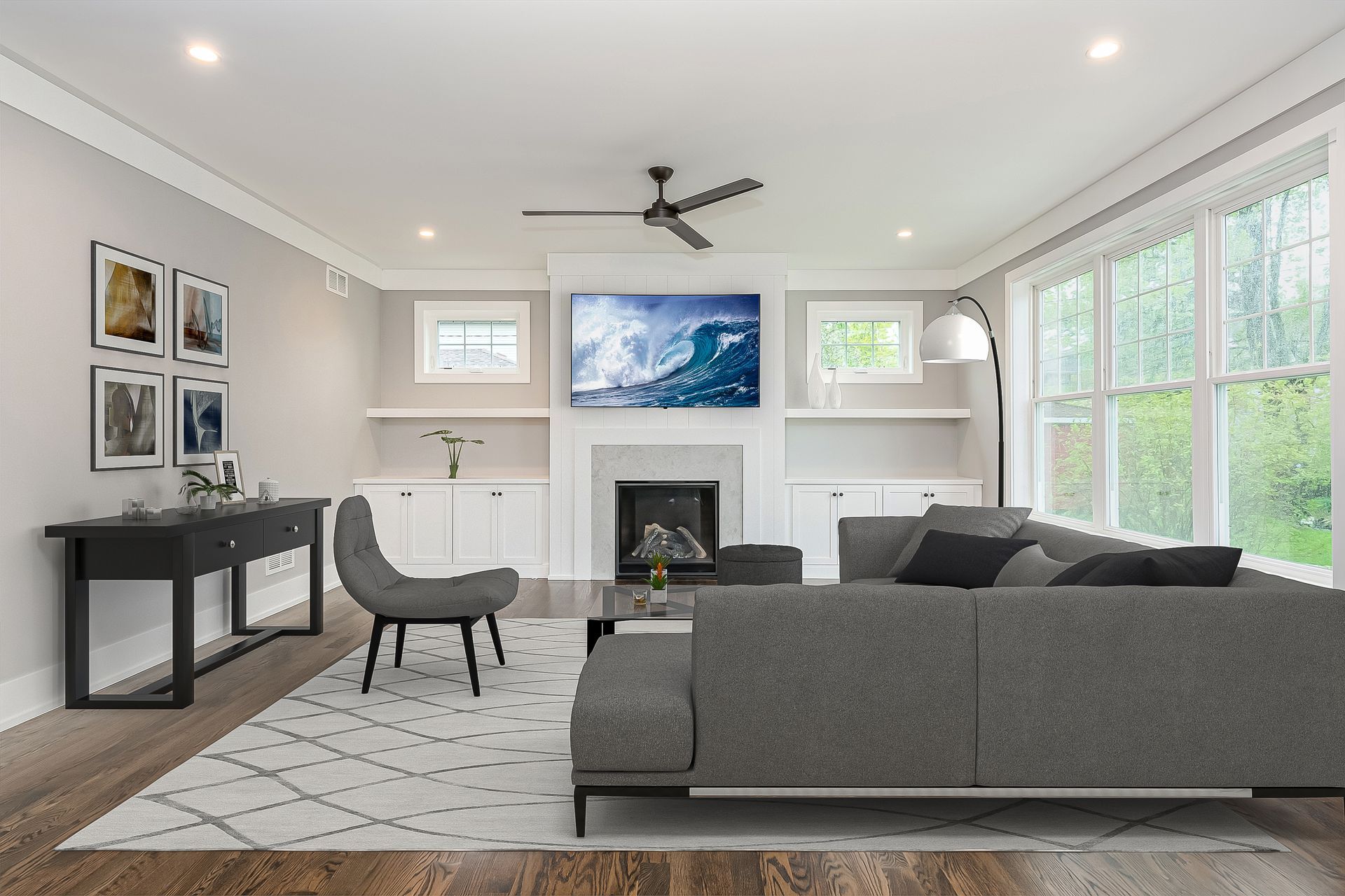 Living room with grey sofa, fireplace, TV, large windows, art, and wood floors.