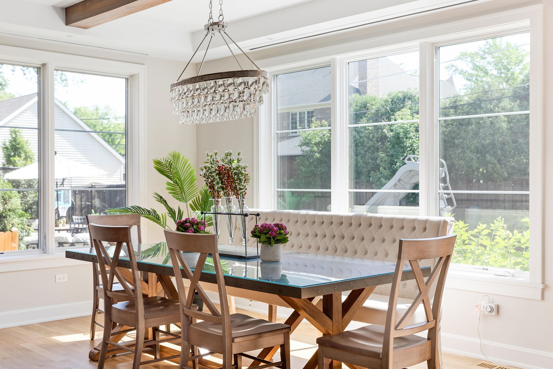Dining room with wooden table and chairs, a tufted bench, and a crystal chandelier by large windows.