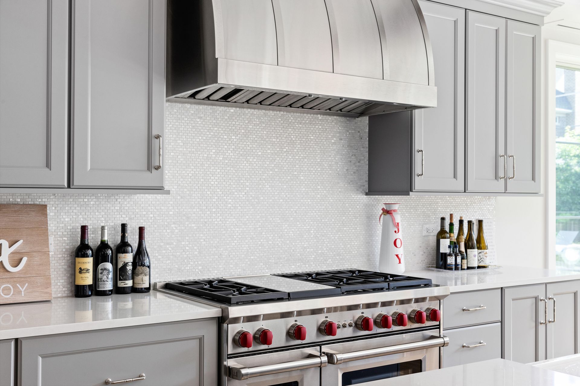 Gray kitchen with stainless steel stove, cabinets, and range hood. Backsplash is white, wine bottles on countertop.