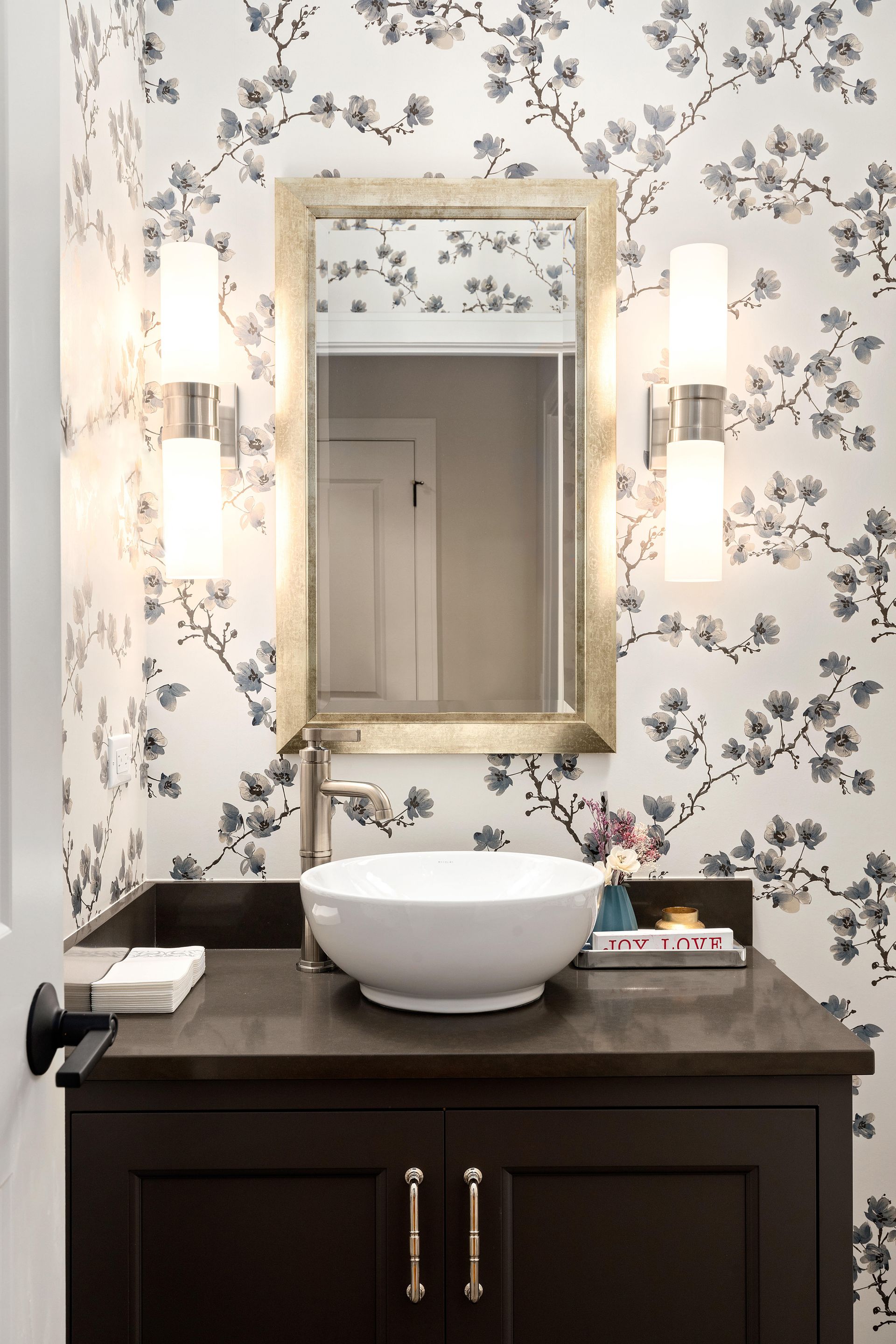 Bathroom vanity with a white bowl sink, dark cabinet, floral wallpaper, and a gold-framed mirror.