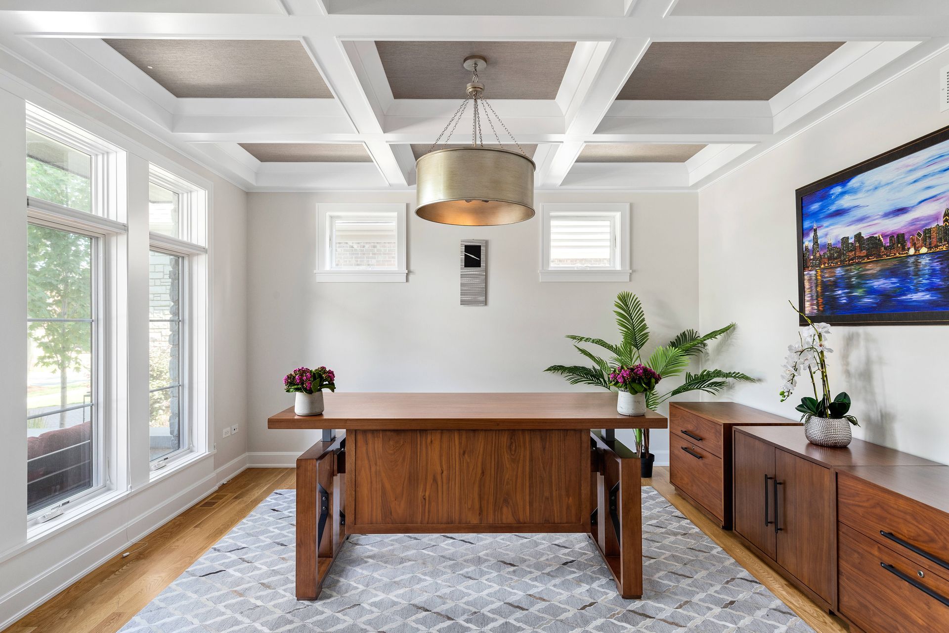 A home office with a wooden desk, cabinet, and a cityscape painting, with a light gray rug, and a coffered ceiling.