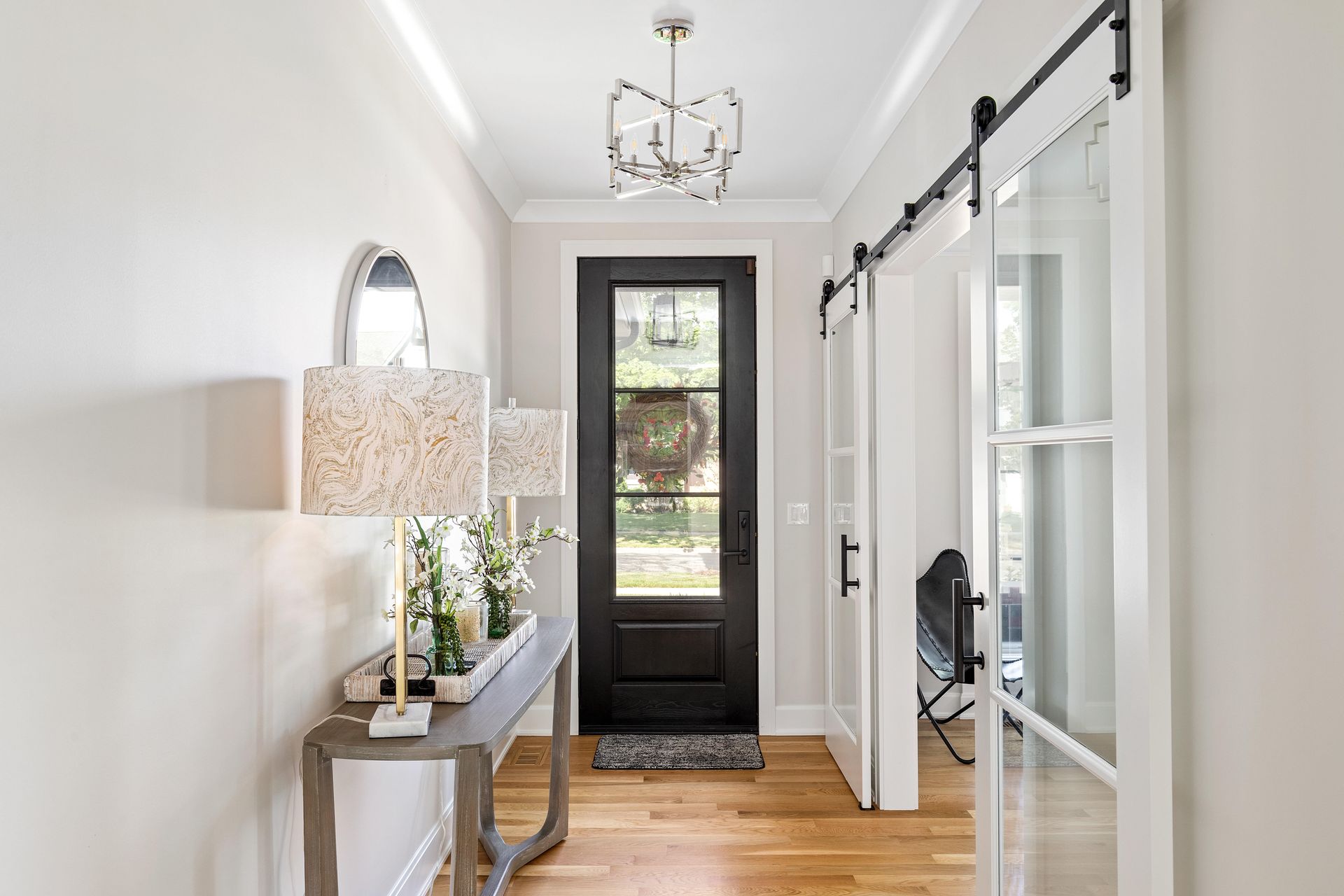 Hallway with light walls, wood floor, table with lamps, black door with window, and sliding glass doors.