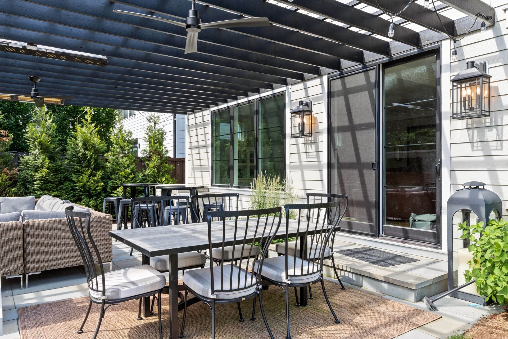 Patio with dining table, chairs, and sofa under a black pergola next to a white house.