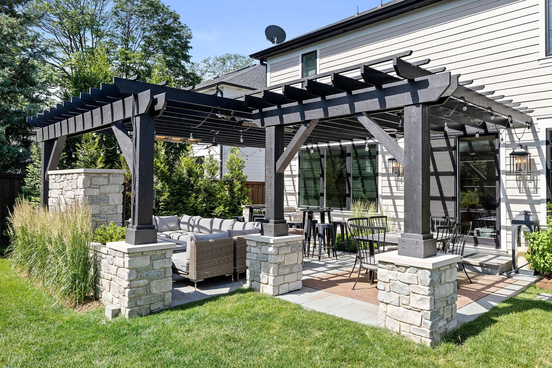 Black pergola over patio with stone pillars and outdoor furniture, beside a two-story house.