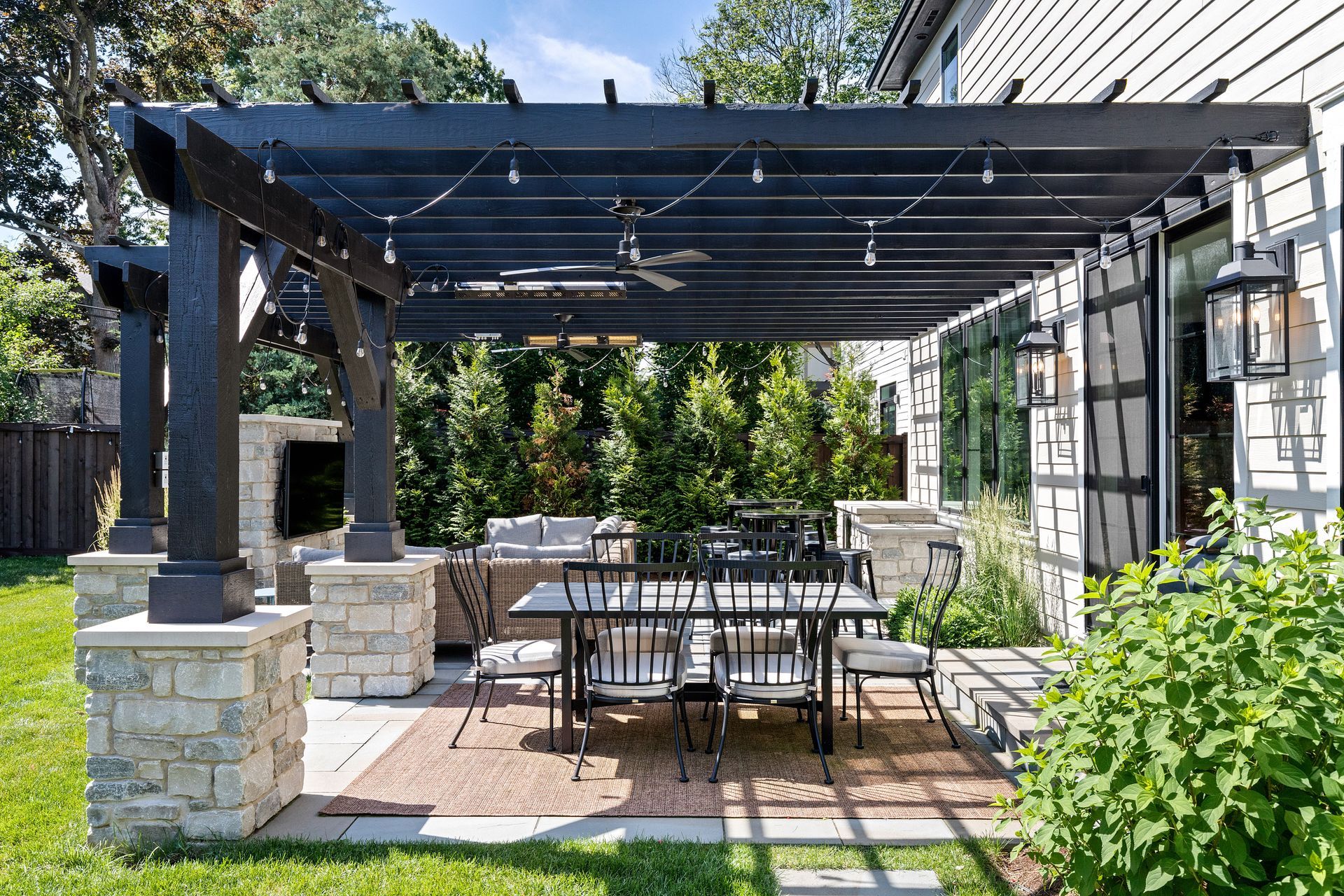 Black pergola over outdoor dining area with lights, adjacent to a white house with stone accents.