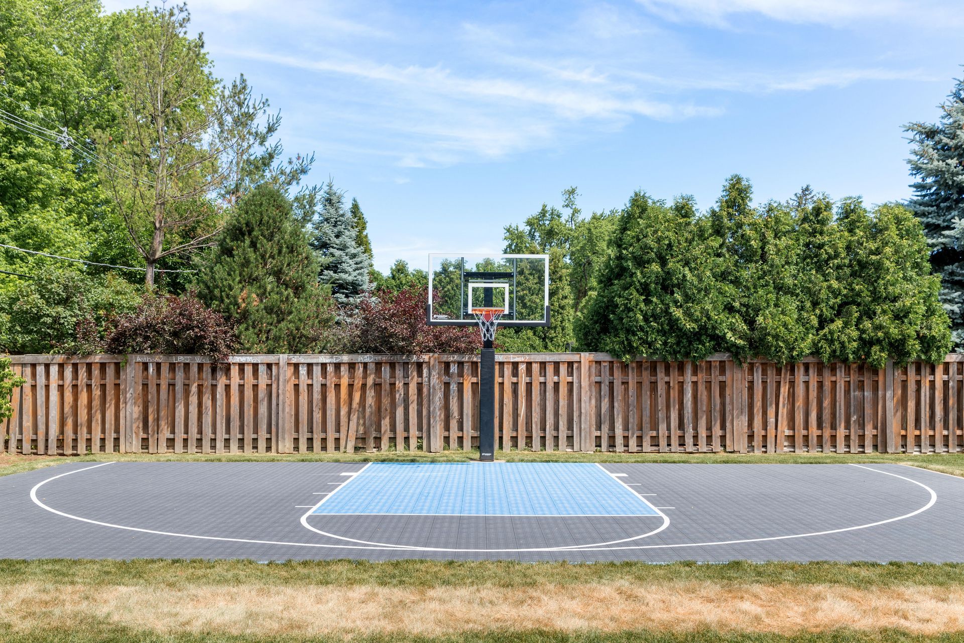 Basketball court in a backyard, with a hoop, blue and gray court, and wooden fence.