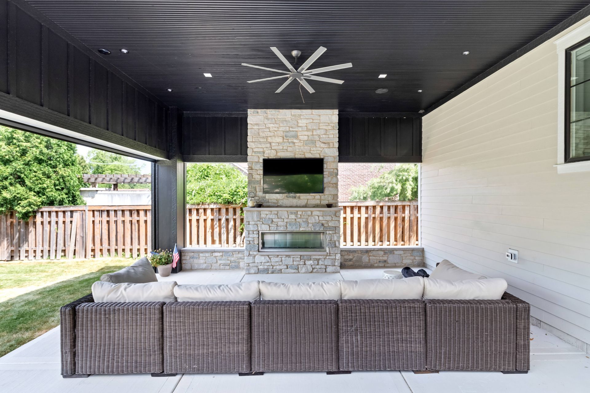 Covered outdoor living space with fireplace, TV, sectional seating, and ceiling fan.