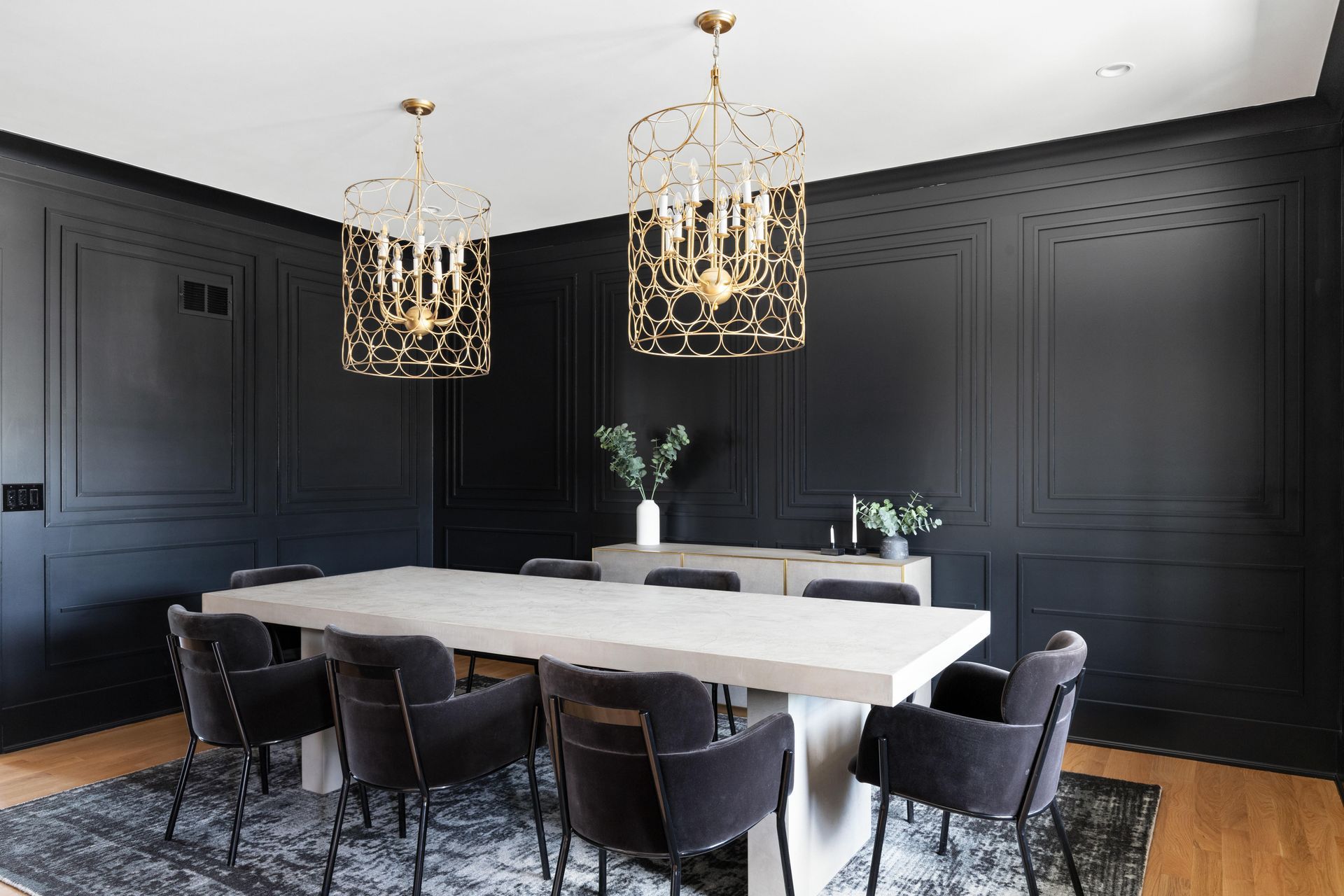 Dining room with black paneled walls, a long white table, dark chairs, and gold chandeliers.