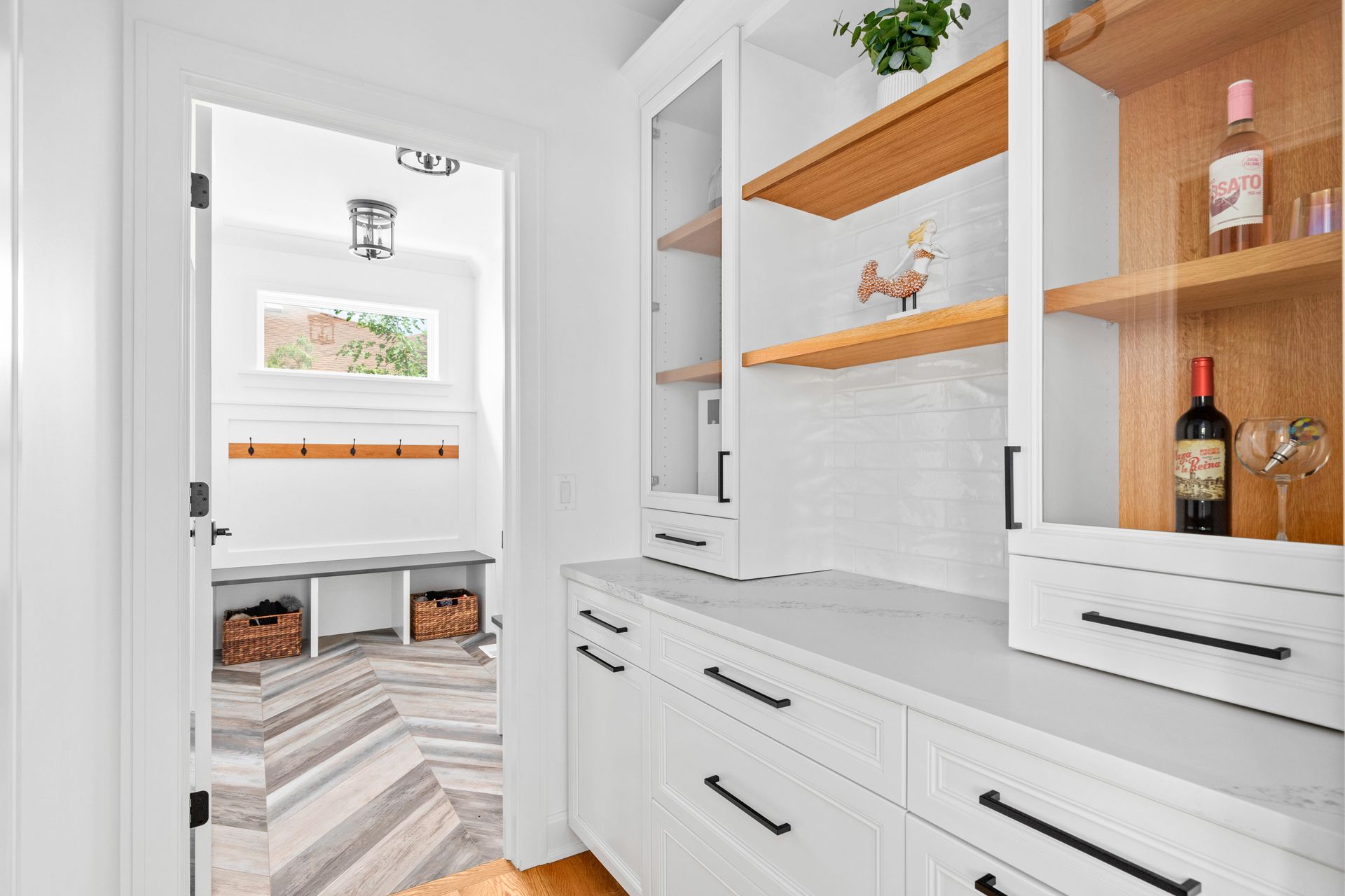 White pantry with open cabinets, wine, and a view of the entryway with wood herringbone floors.