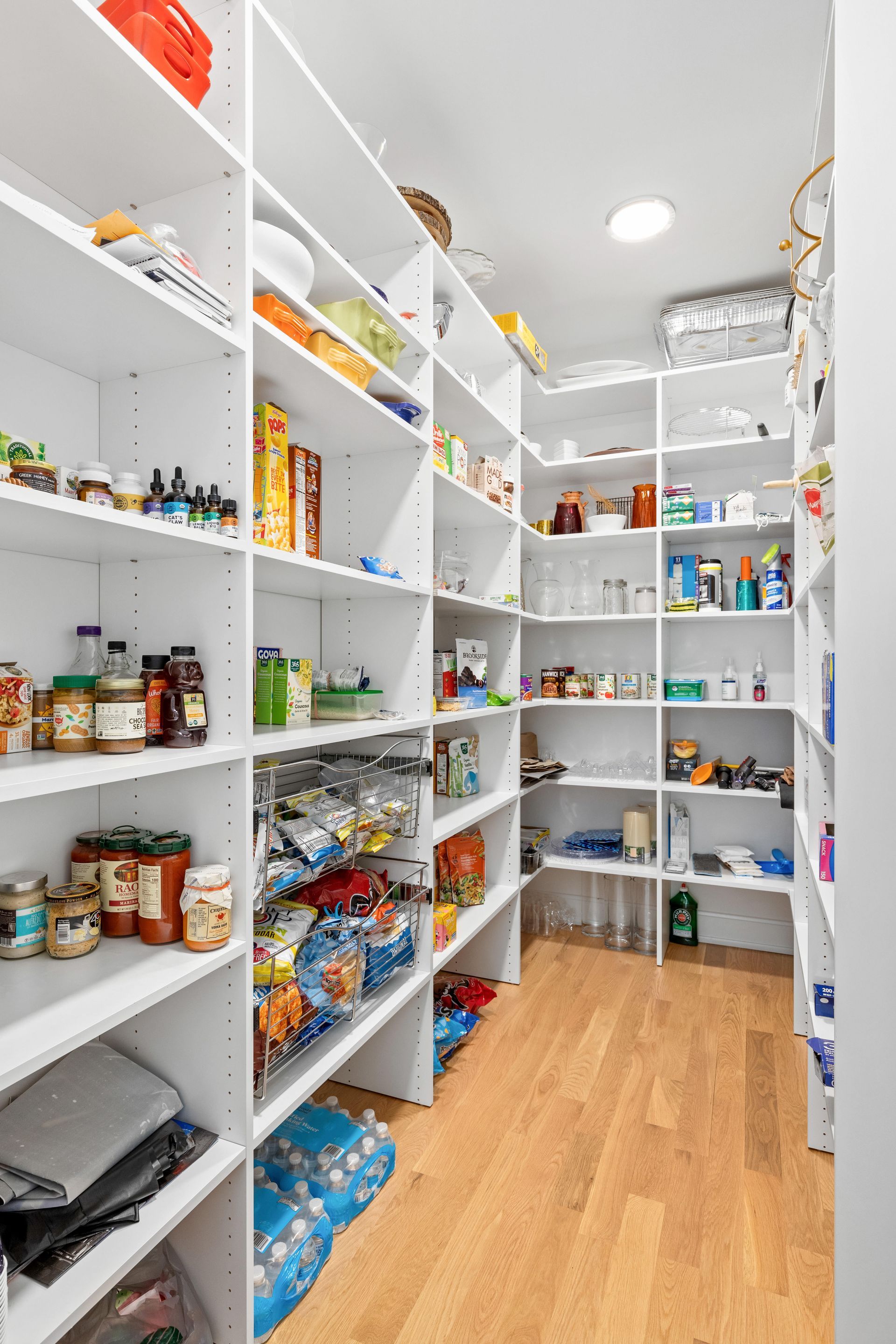 Walk-in pantry with white shelves filled with food and supplies, wooden floor.