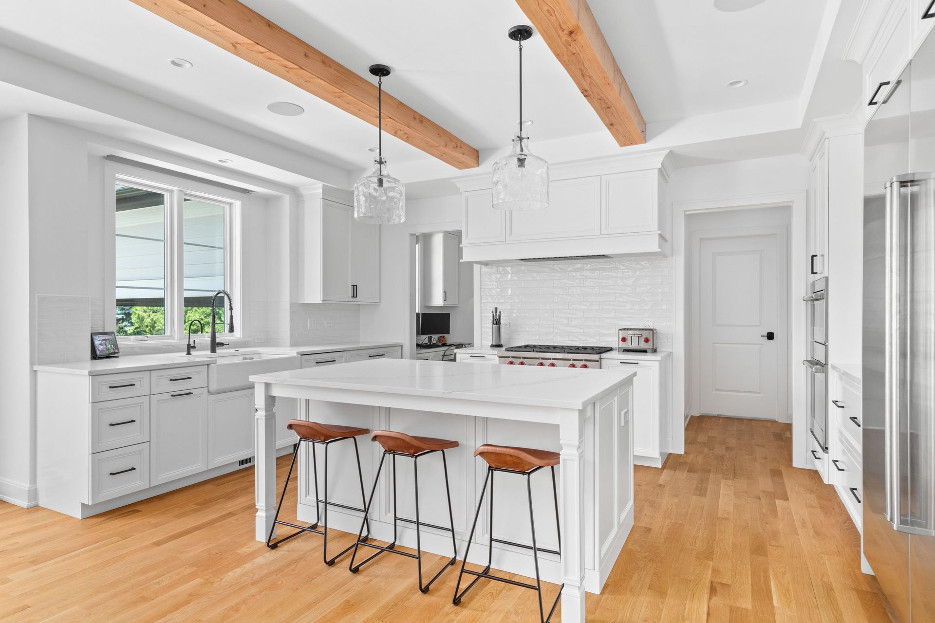 White kitchen with island, wood beams, and hardwood floors. Three bar stools at island.