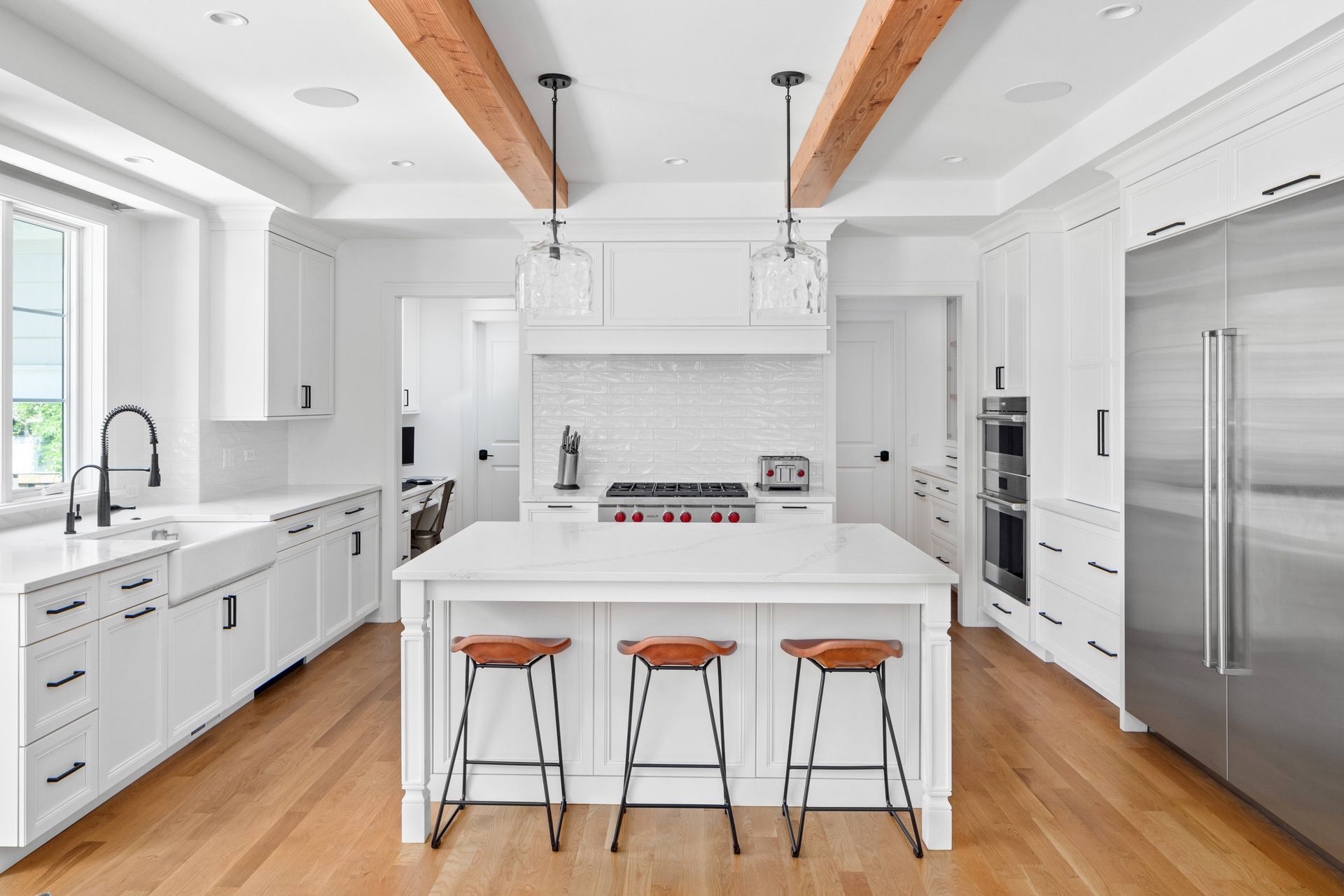 White kitchen with wood beams, island with stools, and stainless steel appliances.