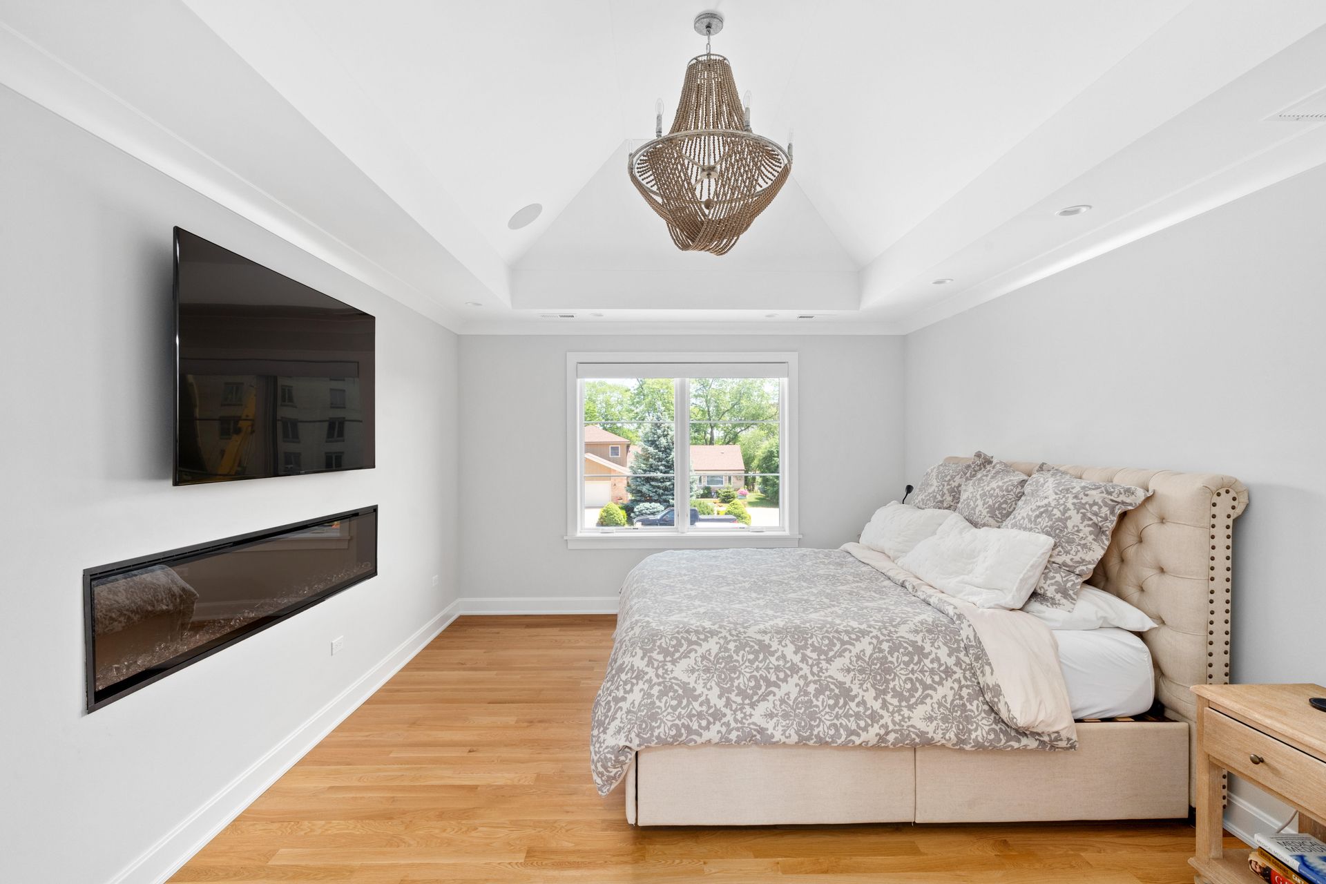 Bedroom with bed, TV, window, and chandelier. Light wood floor, white walls, and gray patterned bedding.
