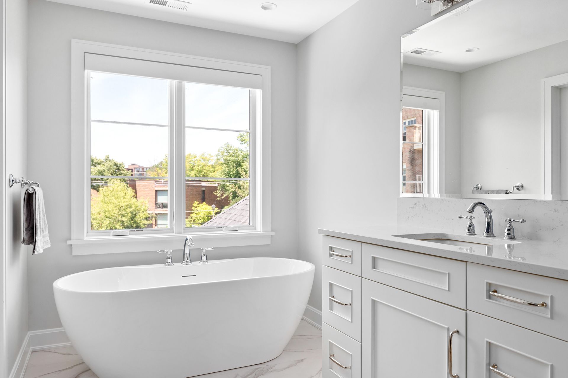 Modern white bathroom with freestanding tub, vanity, and window overlooking greenery.