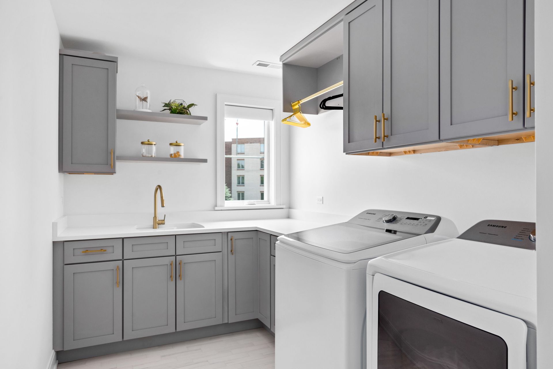 A laundry room with gray cabinets, a white sink and appliances, gold hardware, and a window.