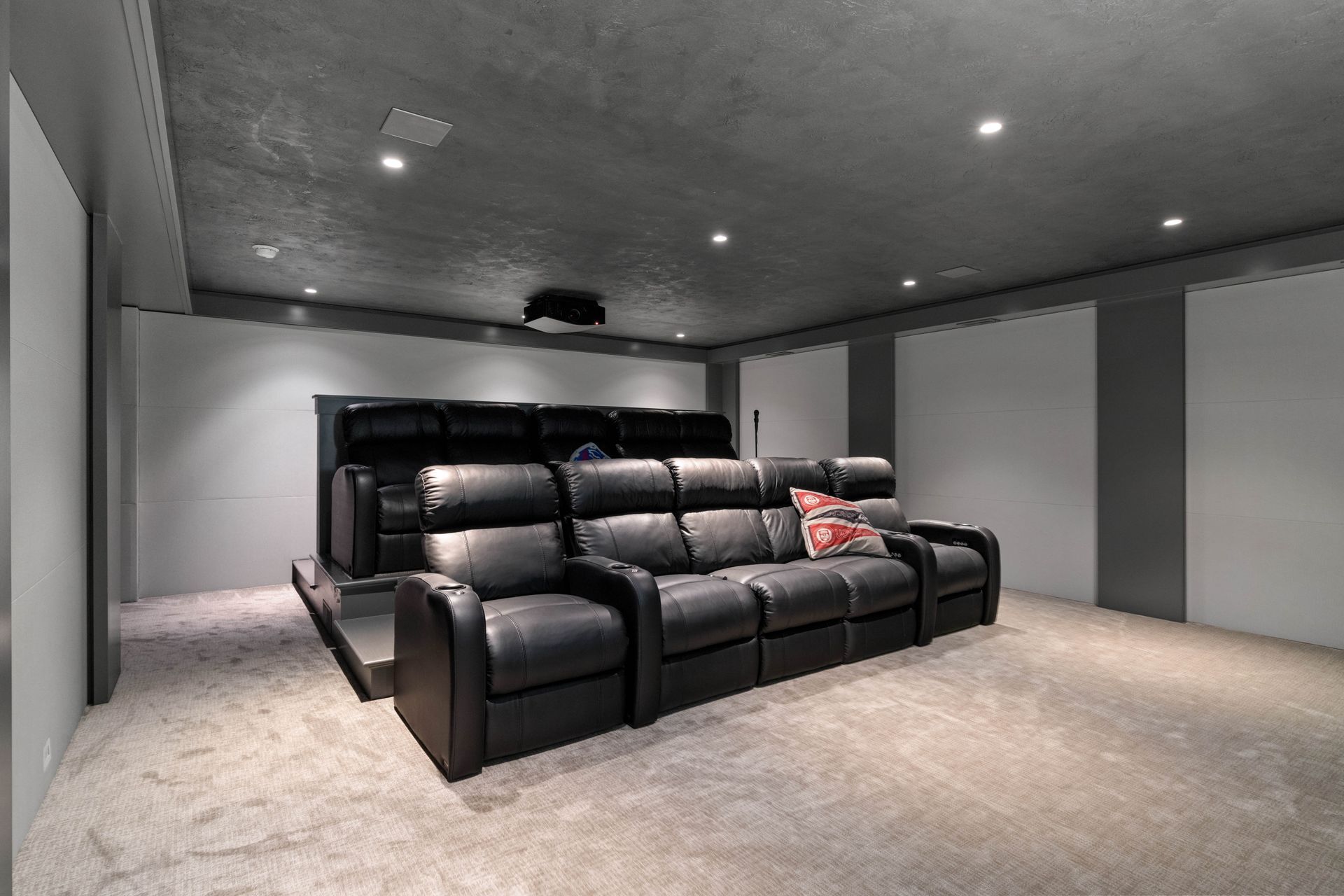 Home theater with tiered black leather recliners, projector, gray walls, and light carpet.