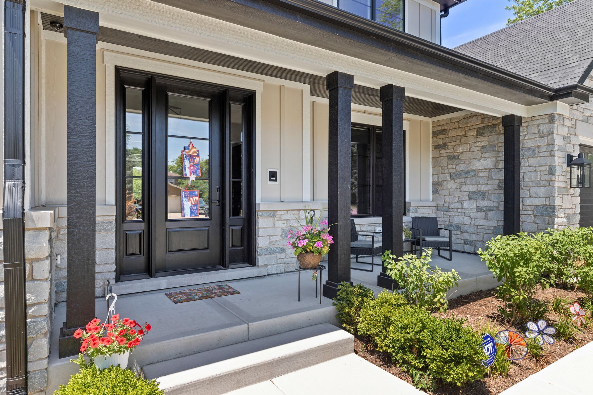 Front porch with black door and pillars, stone and light siding, and flowers.