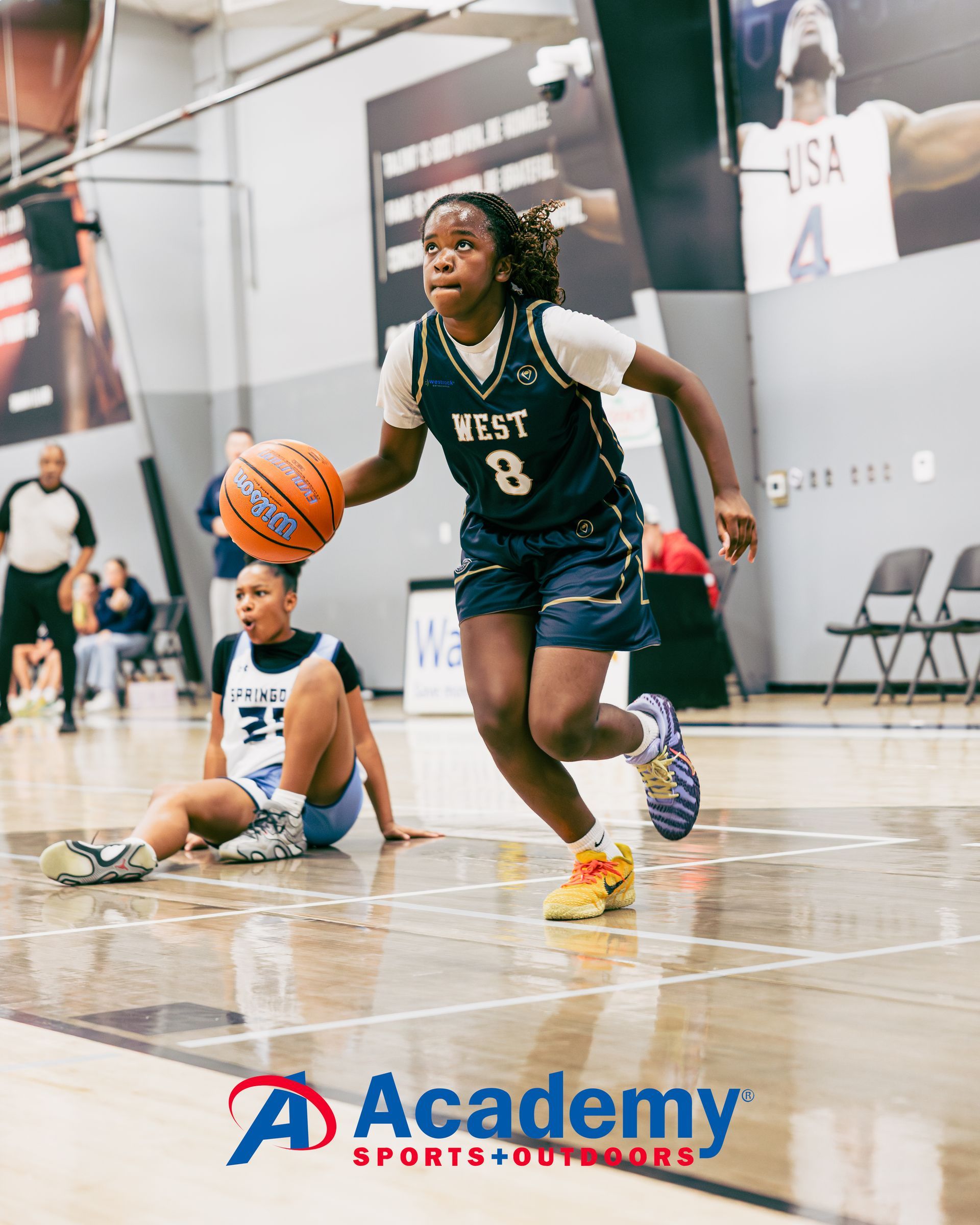 Basketball player dribbling ball, another player on the floor. Academy Sports logo in the foreground. Indoor court setting.