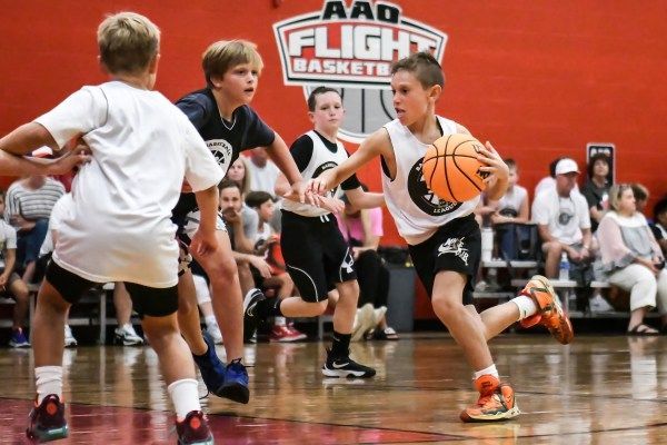 Boys playing basketball during a game indoors. One dribbles orange ball; others defend.