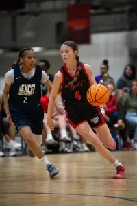 Basketball game: Player dribbles toward the basket, followed by a defender. Indoor setting.