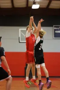Two young basketball players jumping to shoot the ball. One in red, one in black. Indoor court.