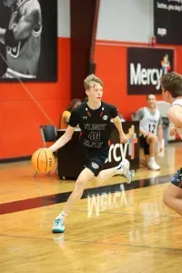 A young basketball player dribbles the ball on a court. He's wearing a black jersey and is mid-stride.