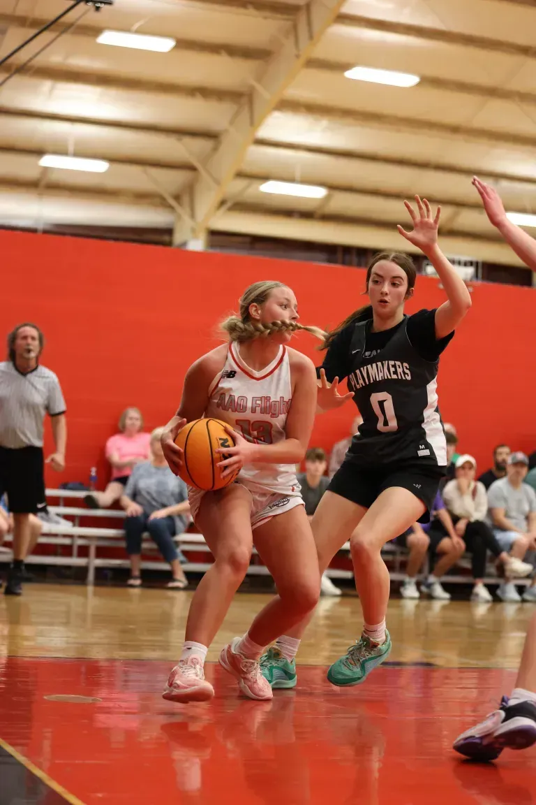 Basketball player in red dribbles past a defender in black, spectators in background.