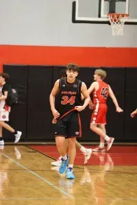 Basketball player, number 34, in black uniform, looks forward in a gym, others blurred in the background.