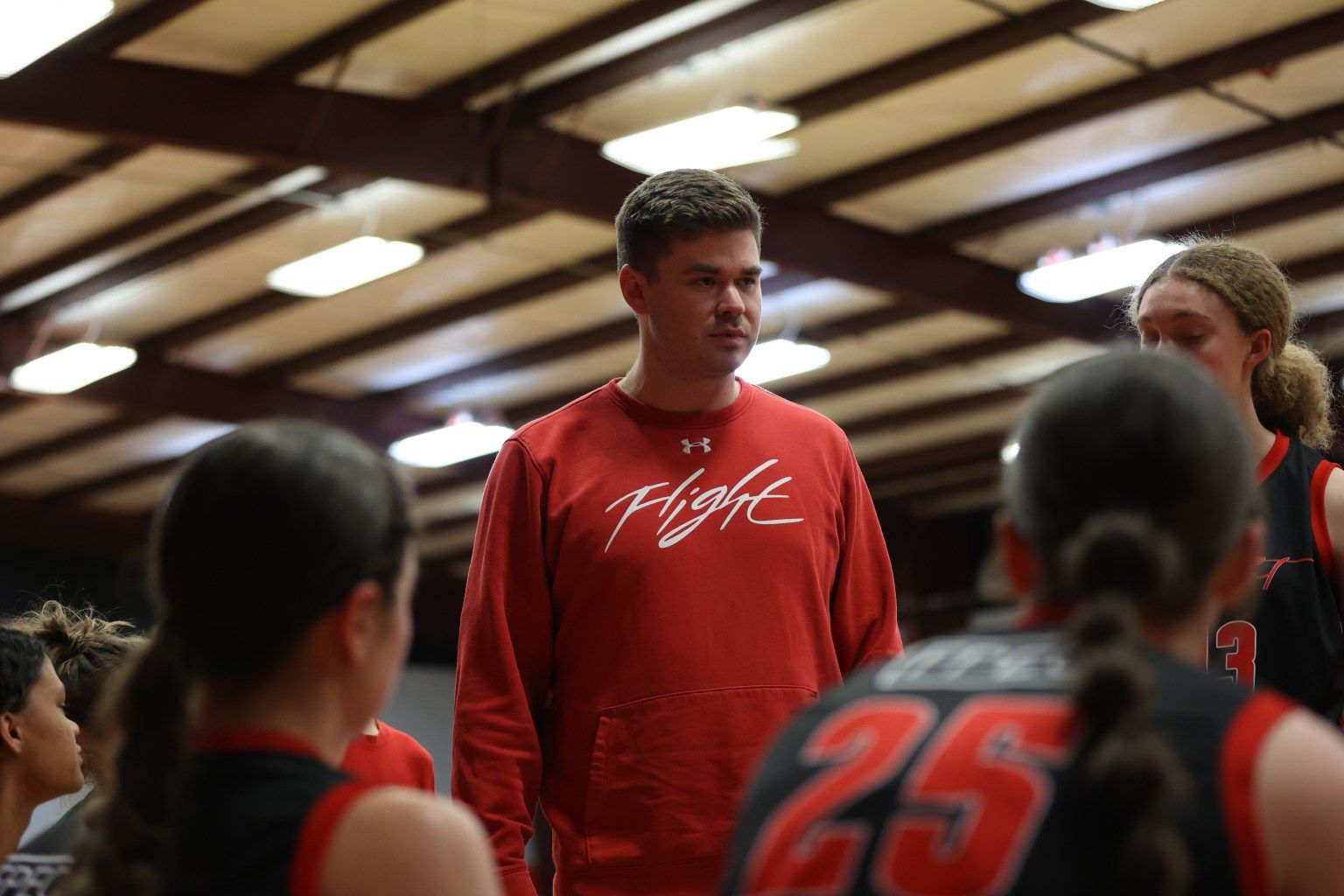 Basketball coach in red sweatshirt talking to a huddle of players on a court.