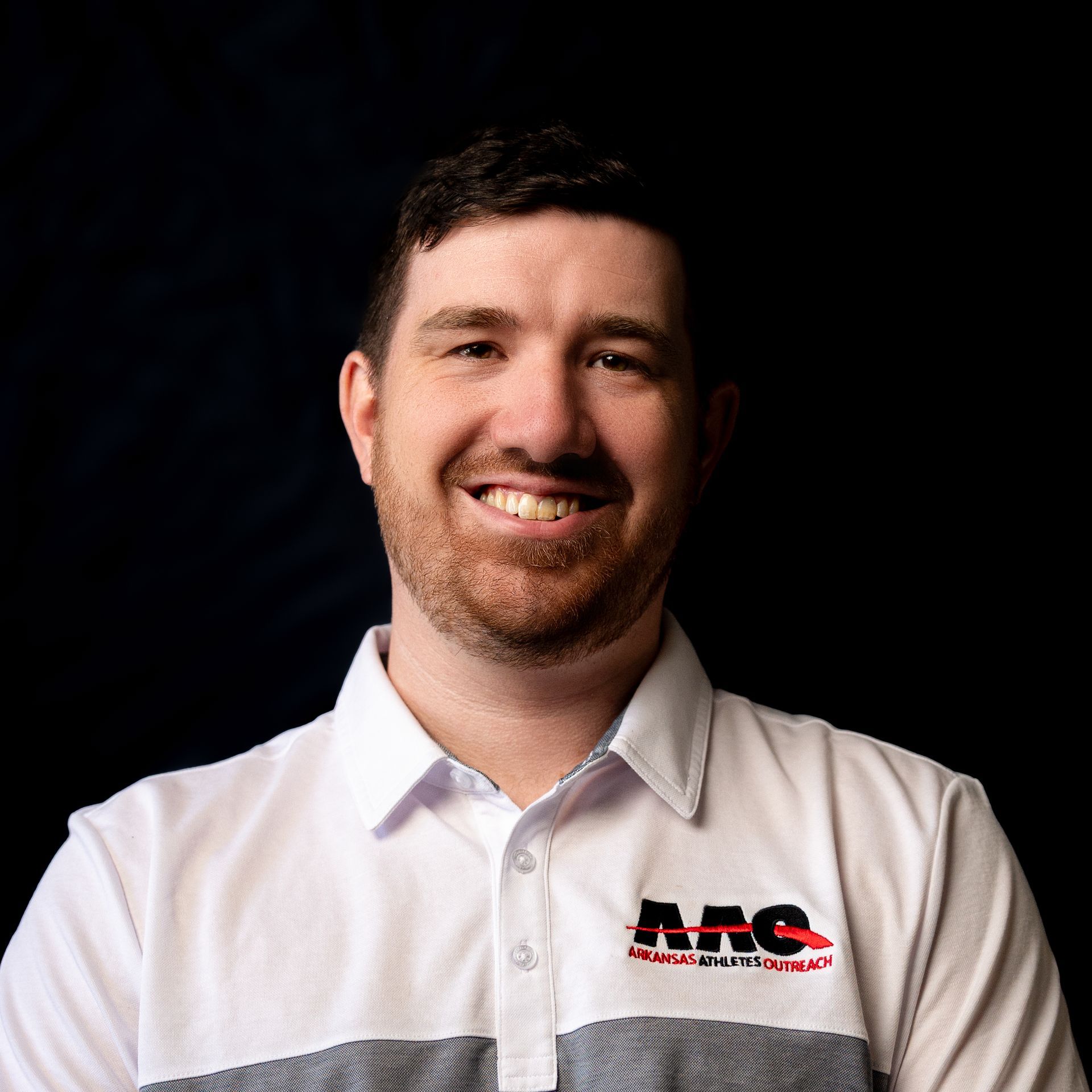 Smiling man in a white polo shirt with a logo, against a dark background.