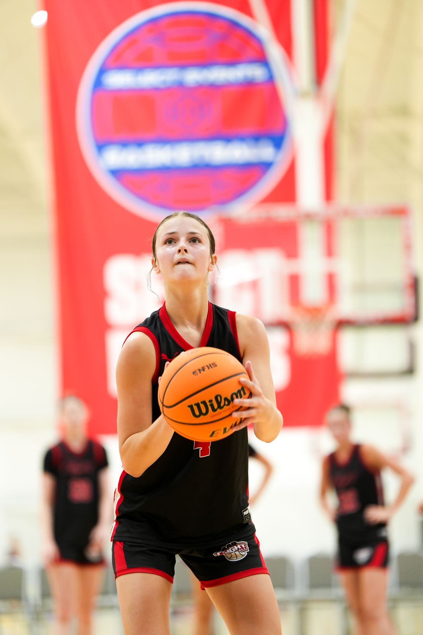 Woman in black basketball uniform shooting a free throw; indoors, others in background.